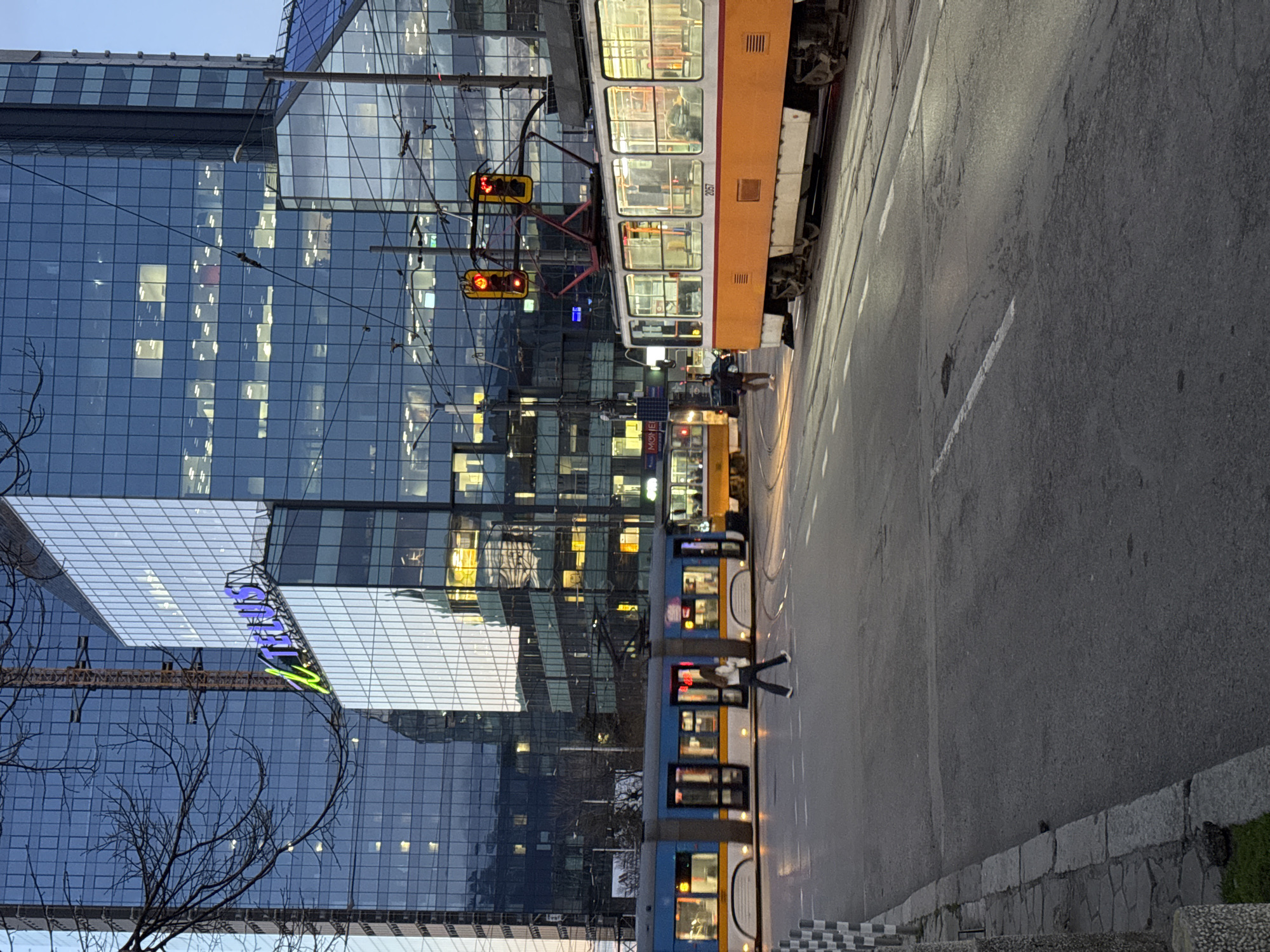 The image shows a street in Sofia, Bulgaria, with a building in the background and two trams.
The building in the background is a modern, high-rise building with a glass facade. The trams are both running on the same track, one is yellow and the other is blue. The street is a paved road with a sidewalk on the right side. There is one person walking along the street. There is a tall metal structure near the buildings.