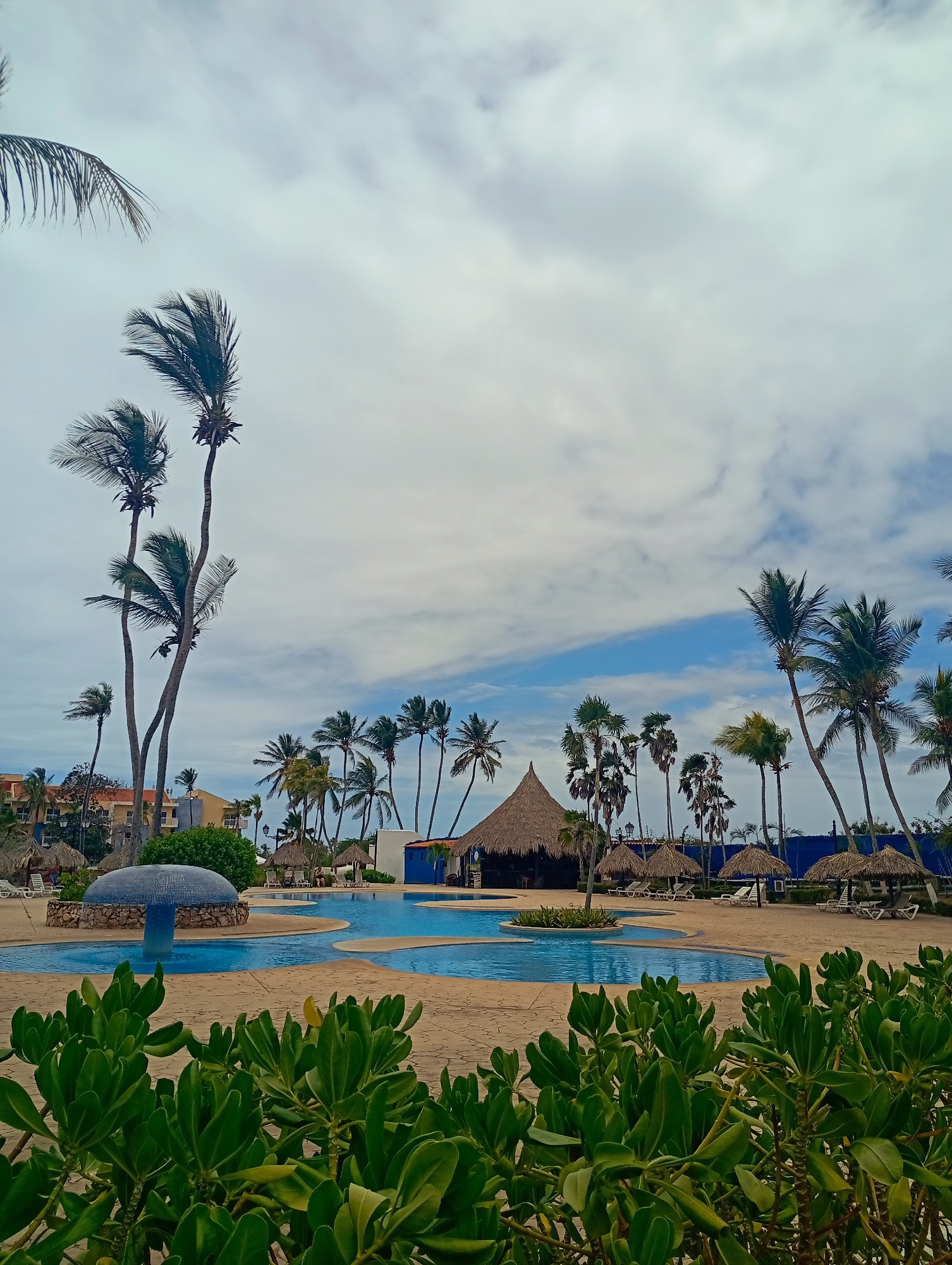 The image shows a resort pool area in Playa El Agua, Venezuela. The pool is multi-tiered and surrounded by tan-colored paving. In the foreground are green bushes, partially obscuring the view. Palm trees are scattered throughout the scene, with some leaning due to the wind. Several thatched-roof structures, including small umbrellas and a larger bar area, are present. In the background, buildings with orange roofs are visible. The sky is partly cloudy, with patches of blue sky breaking through. No people are visible in the scene.