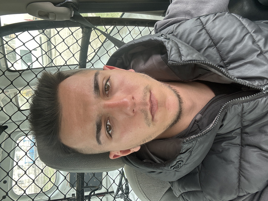 This image depicts a male individual within a vehicle in Srednia, Bulgaria. The subject, appearing to be a young adult male in his early to mid-twenties, is shown from the chest up, directly facing the camera. He possesses short, dark hair, brown eyes, a straight nose, and faint stubble on his upper lip and chin. He is dressed in a dark grey quilted puffer vest worn over a darker, possibly long-sleeved, garment. The individual is seated in a light grey upholstered vehicle seat. Behind him, a metal mesh security partition is visible, separating the front of the vehicle from a rear compartment. Through the vehicle's windows and the mesh, an exterior scene is discernible, featuring blurred residential or commercial buildings, a white vehicle, and industrial machinery, possibly an excavator with blue elements, under an overcast sky.