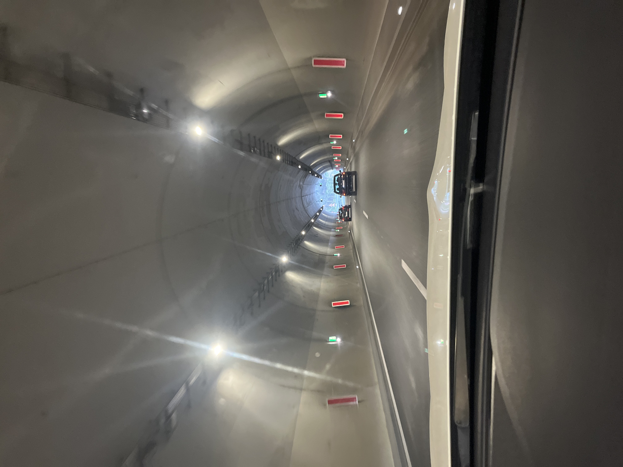 View inside a tunnel with cars in Blagoevgrad, Bulgaria. The tunnel has arched concrete walls with evenly spaced lights and red/green rectangular indicators along the ceiling. Two vehicles are visible in the distance. The image is taken from within a vehicle, with a portion of the vehicle's interior visible on the right side of the frame. The road markings indicate traffic lanes.
