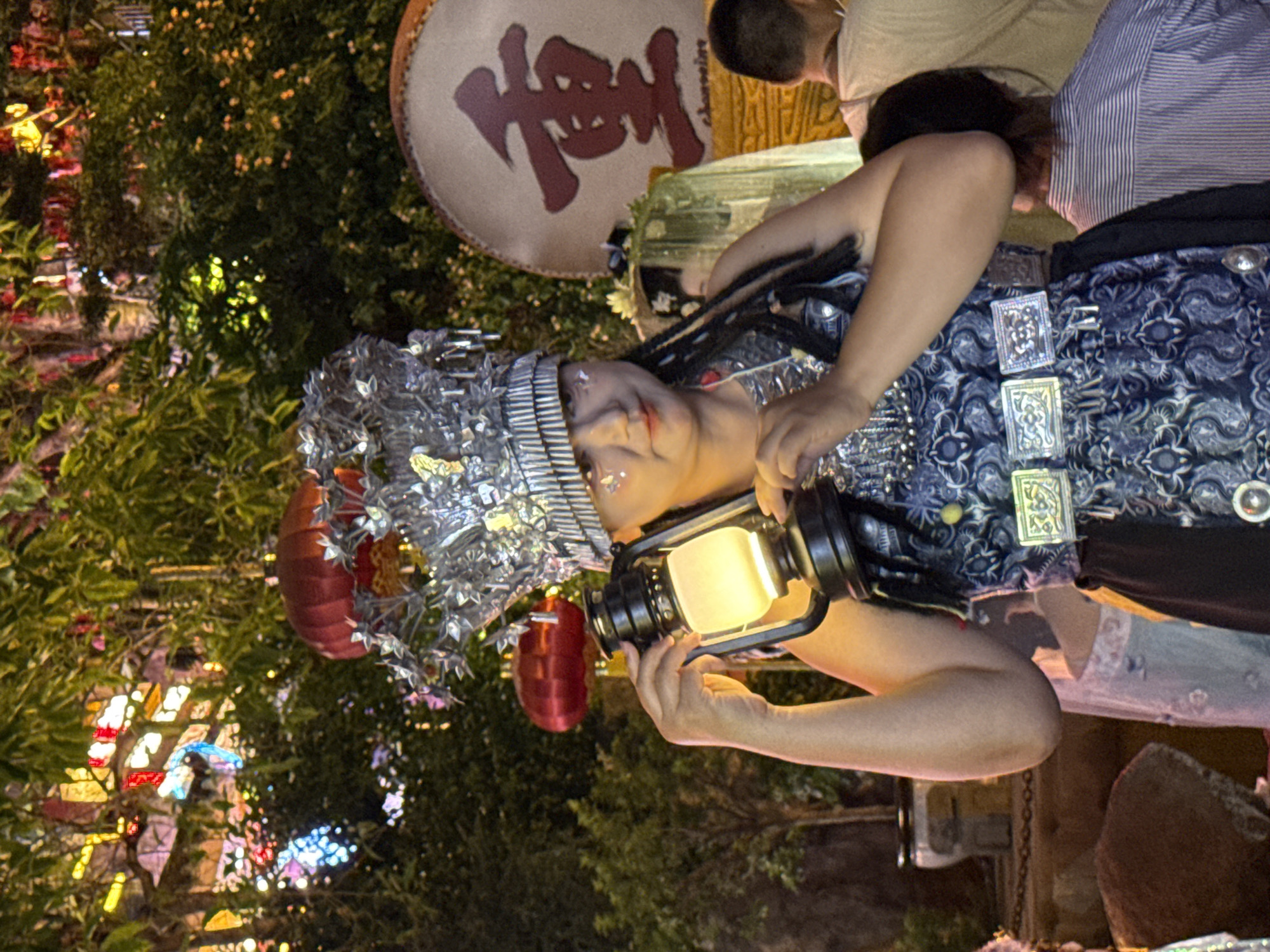 An adult East Asian female, wearing a highly ornate, multi-tiered silver headdress and detailed, patterned blue and silver traditional clothing, holds a lit oil lantern in her right hand and supports it with her left. Her face is adorned with glitter on her cheeks and forehead, and her dark hair is braided. Behind her, to the right, a large circular drum with a prominent Chinese character "重" in red calligraphy is visible, along with the smaller text "Chongqing". The background is dimly lit, with lush green foliage and colorful lights, suggesting an outdoor evening event, possibly in Yuzhong District, Chongqing, China. Other people are visible in the background, out of focus. The overall scene appears festive and cultural.