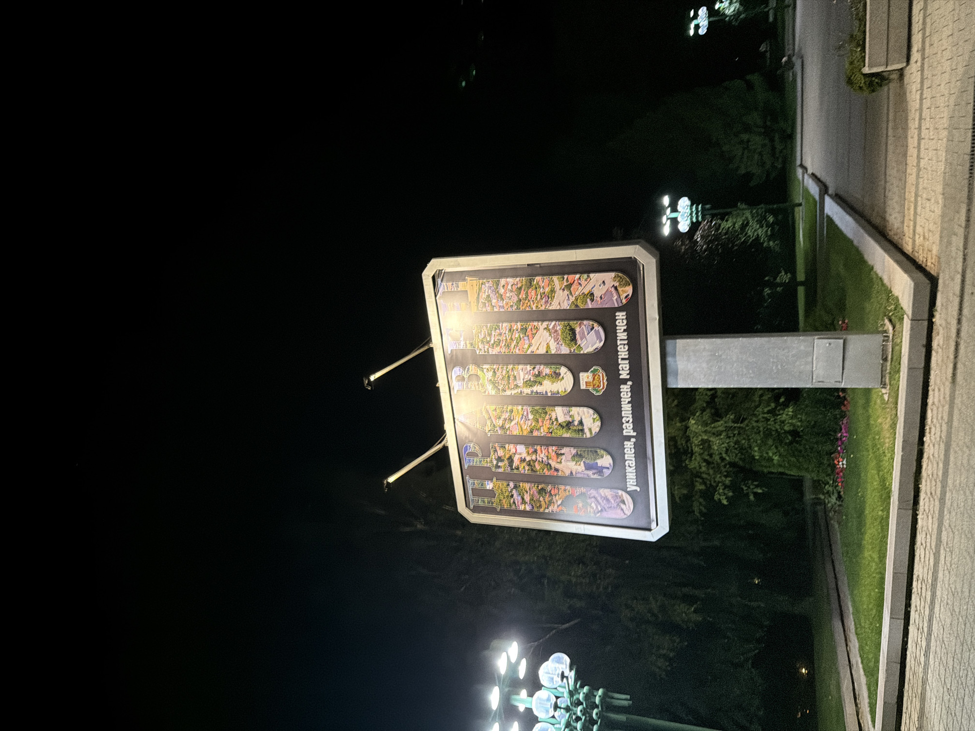 The image shows a night scene in Pravets, Bulgaria. A large billboard is prominently displayed, featuring the word "ПРАВЕЦ" (Pravets) in large letters. Each letter is filled with an aerial view of a town or city. Below the letters, there is text in Bulgarian that reads "уникален, различен, магнетичен" which translates to "unique, different, magnetic". The billboard is illuminated by two spotlights from above. In the foreground, there is a paved walkway and a grassy area. To the left of the billboard, there is a decorative lamppost with multiple lights. In the background, there are trees and more lampposts. The overall impression is that of an advertisement for the town of Pravets.