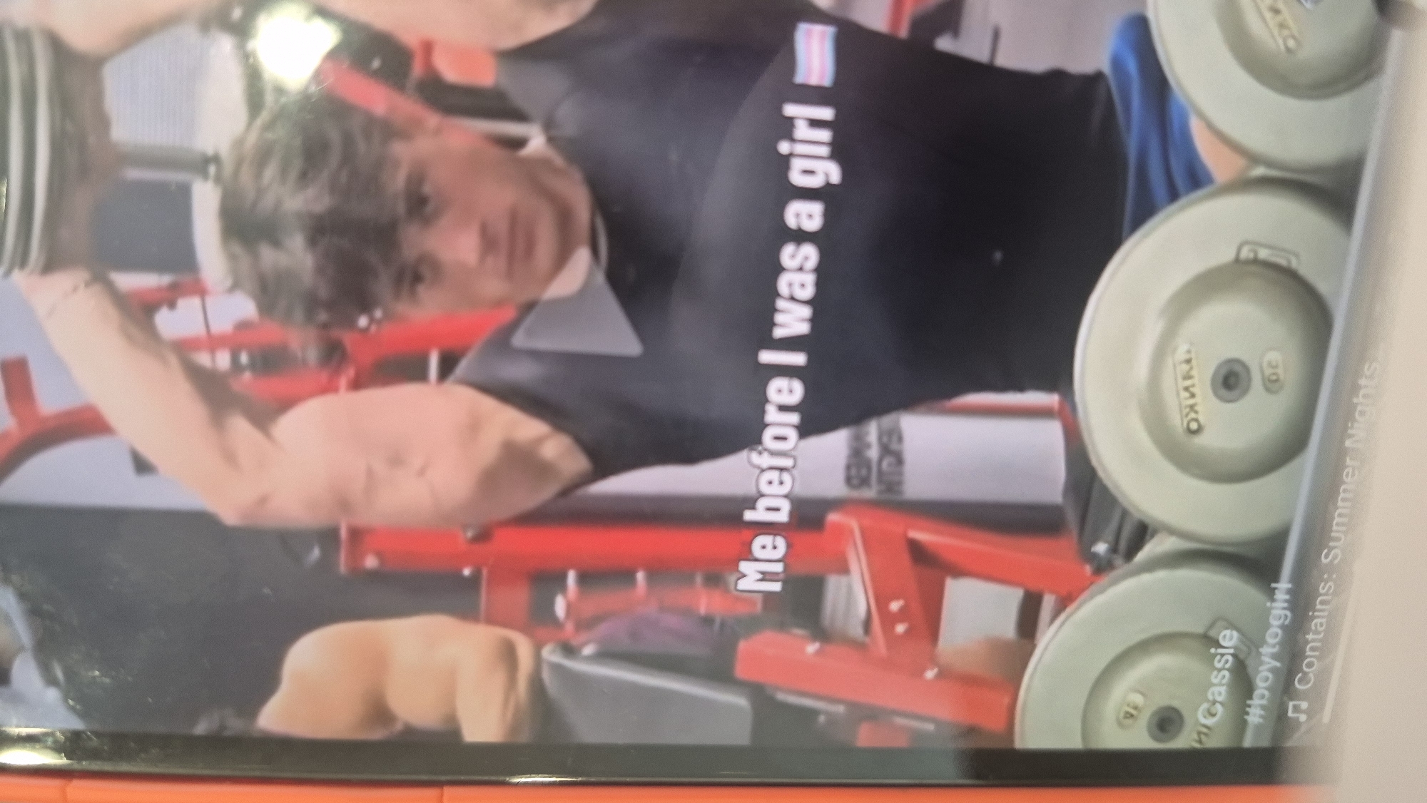 Here's a description of the image:
The image appears to be a screen capture or a photograph of a screen showing a video or image. The primary subject is a person with short brown hair, wearing a dark shirt. They are positioned in what seems to be a gym or fitness area, indicated by the red equipment visible in the background. There are weight plates near the edge of the frame, labeled "20" and "TRWKO".
Text overlay is visible on the image stating, "Me before I was a girl", along with a transgender pride flag. Additional text at the bottom of the screen indicates "#boytogirl," "Cassie" and "Contains: Summer Nights".
No information available in the image suggests the location is Sofia, Bulgaria.
