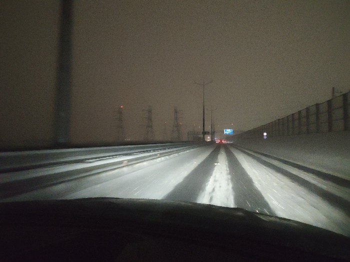The image captures a nighttime, forward-facing view from inside a moving vehicle on a multi-lane highway in Belyaninovo, Russia. The lower portion of the vehicle's dashboard is visible. The road surface is partially covered in snow and ice, with the vehicle's headlights illuminating distinct, wet, clear tire tracks in the immediate foreground and mid-distance. The sky is dark, overcast, and hazy, suggesting snowfall or fog, limiting long-distance visibility.
To the left, a prominent dark vertical structure, possibly a pole or support, stands in the foreground. Further in the background on the left, several large electrical transmission towers, each with a red warning light, are discernible against the dim sky. To the right, a tall, continuous noise barrier or retaining wall, light in color, runs parallel to the highway. A circular blue road sign with a white border is affixed to this wall. Ahead, various streetlights illuminate sections of the road, and an overhead blue road sign with white text is visible. The red tail lights of other vehicles are present in the distance, indicating traffic ahead on the highway. No people are visible.