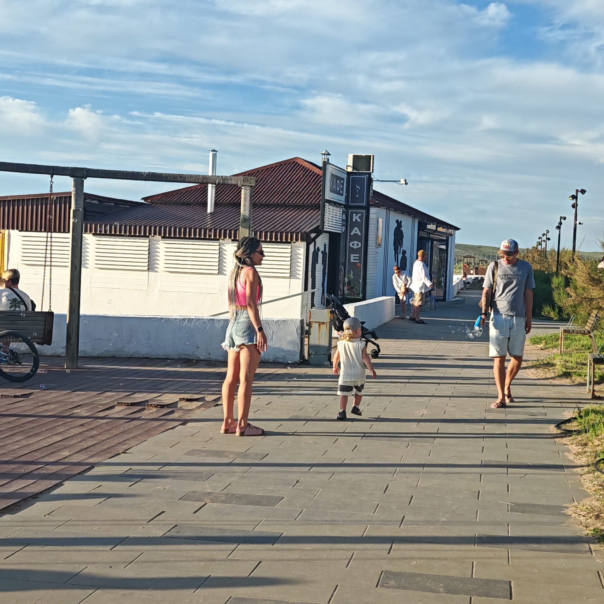The image depicts an outdoor scene on a sunny day in городское поселение Щелкино, Ukraine, focusing on a promenade adjacent to a building. At least six individuals are present.
In the center-left foreground, a woman with long dark braids, sunglasses, a pink crop top, and denim shorts stands facing right. To her right, a small child, likely a toddler, wearing a light top and striped shorts, walks ahead in the same direction. Behind the child, a dark, compact stroller is visible near the building's entrance.
Further to the right, a man wearing a grey t-shirt, light-colored shorts, a baseball cap, and sunglasses walks forward, slightly angled towards the left. He holds a blue bubble wand, and several clear, spherical bubbles are floating in the air around him.
To the far left, a person wearing a light shirt with a palm tree pattern is seated on a wooden swing bench. A bicycle wheel is partially visible near the swing.
In the background, two men are positioned near the entrance of the main building. They are both wearing light-colored long-sleeved shirts and shorts, engaged in casual standing or walking. Further right and in the background, another individual in a white long-sleeved shirt walks away from the scene.
The building in the mid-ground features a reddish-brown corrugated metal roof and white walls with horizontal wooden shutters on the windows. A prominent sign on its facade displays "КАФЕ" in Cyrillic text, vertically and horizontally. To the right of this sign, artistic black silhouettes of human figures are painted on the white wall. Multiple metal chimneys are visible on the roof.
The ground consists of two distinct sections: a darker, possibly wooden or composite boardwalk on the left, and a lighter grey paved pathway made of rectangular pavers with thin groove lines on the right. Long shadows are cast across the pathway, indicating a low sun angle, suggesting either early morning or late afternoon.
Along the right side of the paved path, multiple tall, dark streetlights with conical fixtures extend into the distance. Sparse, dry vegetation, including grass and low shrubs, borders the pathway on the right, and a wooden slat bench is visible among the greenery in the far right. The sky overhead is bright blue with scattered, wispy white clouds. In the far background, a gentle, treeless hill rises with sparse ground cover.