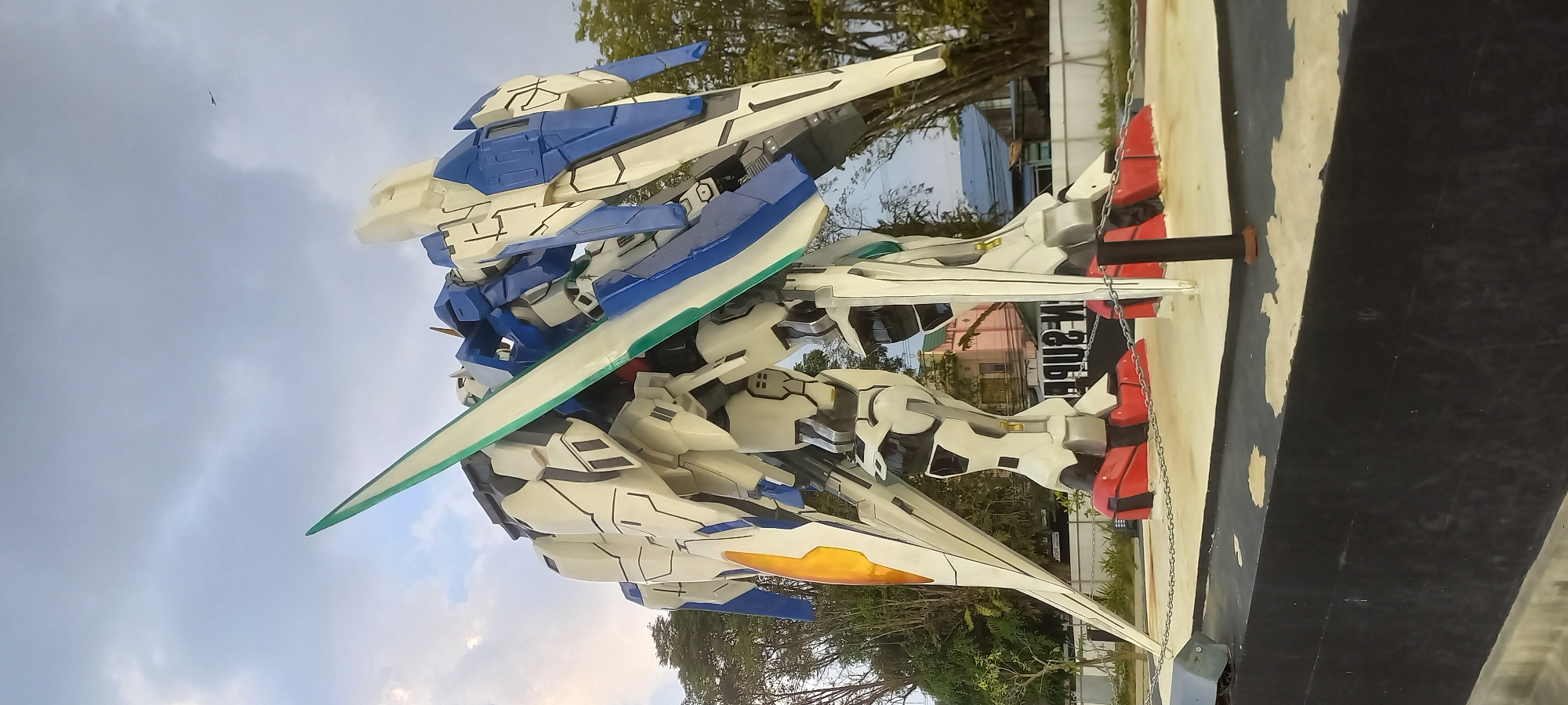 A large, white, blue, and yellow statue of a mecha-style robot stands outdoors. The robot is in a dynamic pose, with one leg forward and a large sword held diagonally across its body. The robot is on a raised platform, surrounded by a chain fence. In the background, there are trees, houses, and a cloudy sky. A sign with text is visible behind the robot. The overall scene appears to be in a public space, possibly a park or plaza.