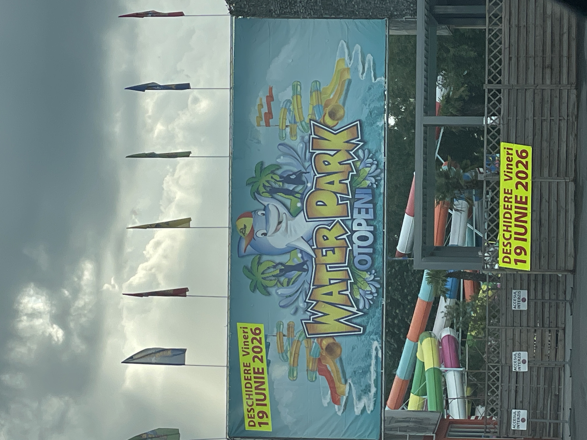 The image shows an advertisement for the Water Park Otopeni in Otopeni, Romania. The advertisement features a cartoon shark and mentions the opening date as Friday, June 19, 2026. Several colorful water slides are visible behind the sign. There are also a series of colorful flags on poles and access signs that read "ACCESUL INTERZIS." The sky is cloudy.