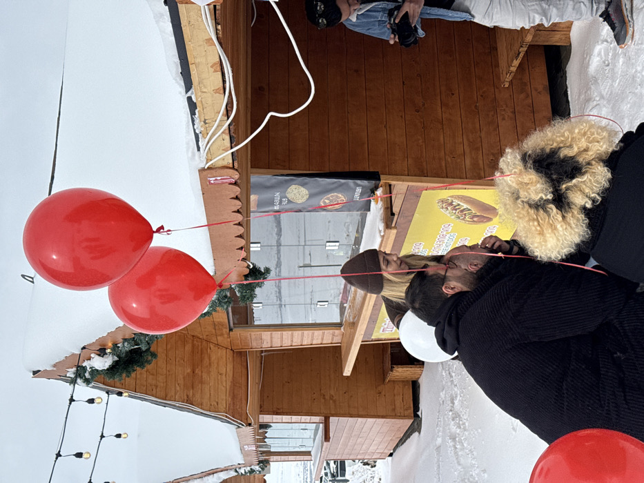 This image captures a winter scene in Yeghipatrush, Armenia, featuring a group of individuals gathered around wooden kiosks on snow-covered ground. The sky is overcast and white.
In the foreground, two large, glossy red balloons float upwards, tethered by red ribbons, one ribbon extending towards a person in the foreground. A white balloon is partially visible behind one of the individuals. The ground is uniformly covered in snow.
Two wooden kiosks with peaked roofs, also covered in snow, are visible. Green garlands are draped beneath the eaves of the kiosks. String lights are strung between the kiosks. The kiosk on the right features a serving counter and a yellow sign displaying "THIN hot dog" and Armenian text, along with an image of a hot dog. Another sign above it displays "MASUN" and "URAN" with graphic elements. White electrical wiring is visible along the roofline of the rightmost kiosk.
Four individuals are present. In the center-left, a male with short dark hair and glasses wears a dark, possibly ribbed, coat and is crouching low, facing right. Behind him and slightly to his left, a female with blonde hair, wearing a brown beanie and a brown jacket, leans forward, looking downwards in the same direction as the crouching male. In the immediate foreground, with their back to the camera, is a person with curly blonde and dark hair, wearing a dark garment, positioned very low. To the right, a male stands, wearing a light blue denim jacket, light-colored pants, and dark sneakers. He holds a black camera, appearing to film or photograph the scene unfolding in front of the kiosk. He has sunglasses perched on his head.