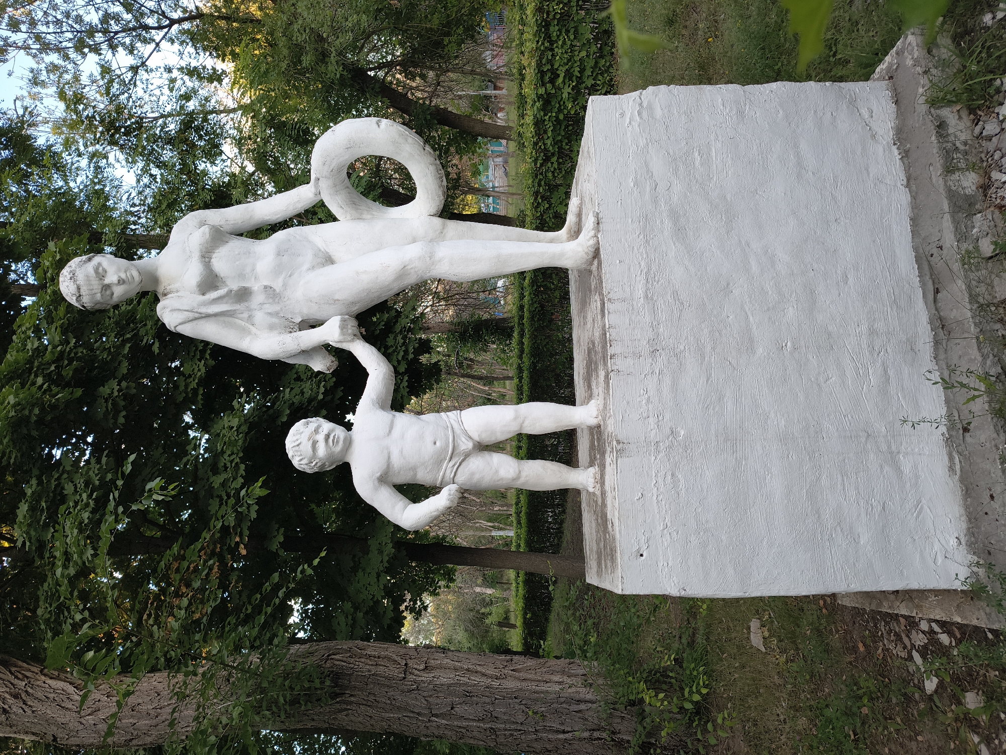 The image shows a white stone sculpture of a woman and a child standing on a white pedestal in Mikhaylovka, Russia. The woman is taller than the child and is holding a life preserver. The child is smaller and is holding the woman's hand. Both figures are nude, and the woman is wearing a cloth draped over her shoulder. The sculpture is situated in a park-like setting with trees and bushes in the background. The lighting suggests it is daytime, possibly late afternoon, with sunlight filtering through the trees. The overall scene appears to be in a public space, likely a park or a recreational area.
