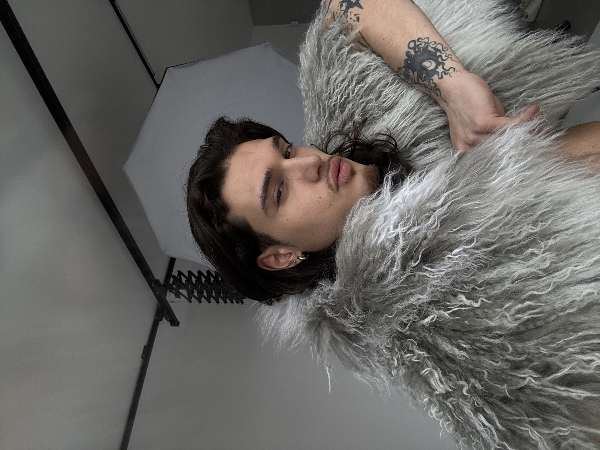 A young adult male subject with dark, wavy hair and light facial hair forming a goatee and mustache poses in an indoor photography studio in Sofia, Bulgaria. He is seen from the chest up, gazing directly at the camera with a slightly narrowed eye and pouted lips. The subject wears a shaggy, light grey faux fur garment that covers his torso and shoulders. His left ear displays a small silver stud in the upper lobe and a silver hoop earring in the lower lobe. A prominent dark tattoo, featuring an organic, multi-tentacled design, is visible on his right forearm, with a smaller star-shaped tattoo partially visible above it on the upper arm. His right hand rests on the fur garment. The background consists of a plain light grey wall. Overhead, black metal track lighting equipment with an accordion-style extension is visible. A large white octagonal softbox, indicative of studio lighting, is positioned in the upper right quadrant of the frame.