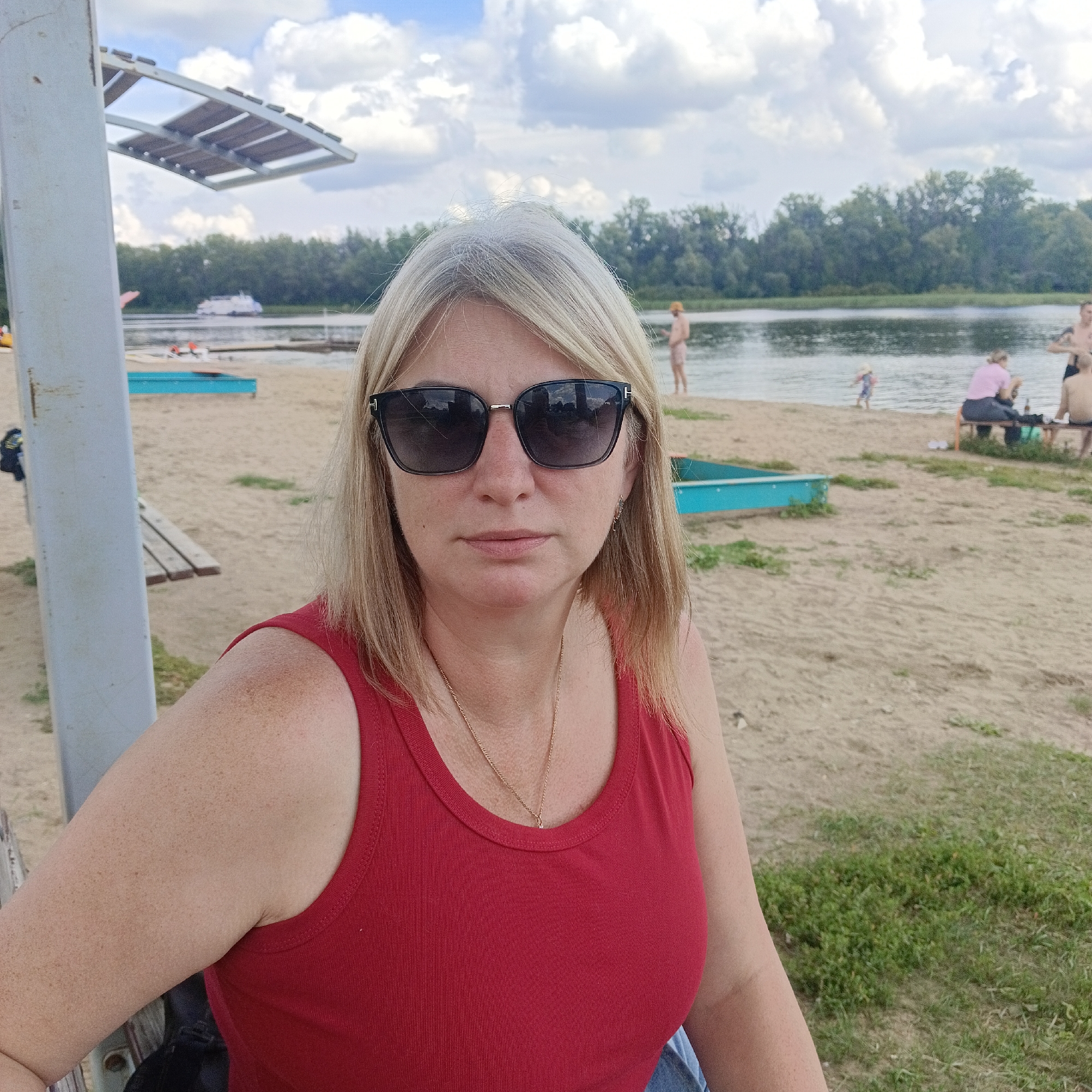 A woman with blonde hair and sunglasses is sitting on a beach in Rozhdestveno, Russia, near a body of water. She is wearing a red tank top and a gold necklace. In the background, there are other people on the beach, some sitting on benches and others near the water. There are also some blue boats on the sand and a white boat in the distance on the water. The sky is cloudy. The overall setting appears to be a summer day at a recreational area.