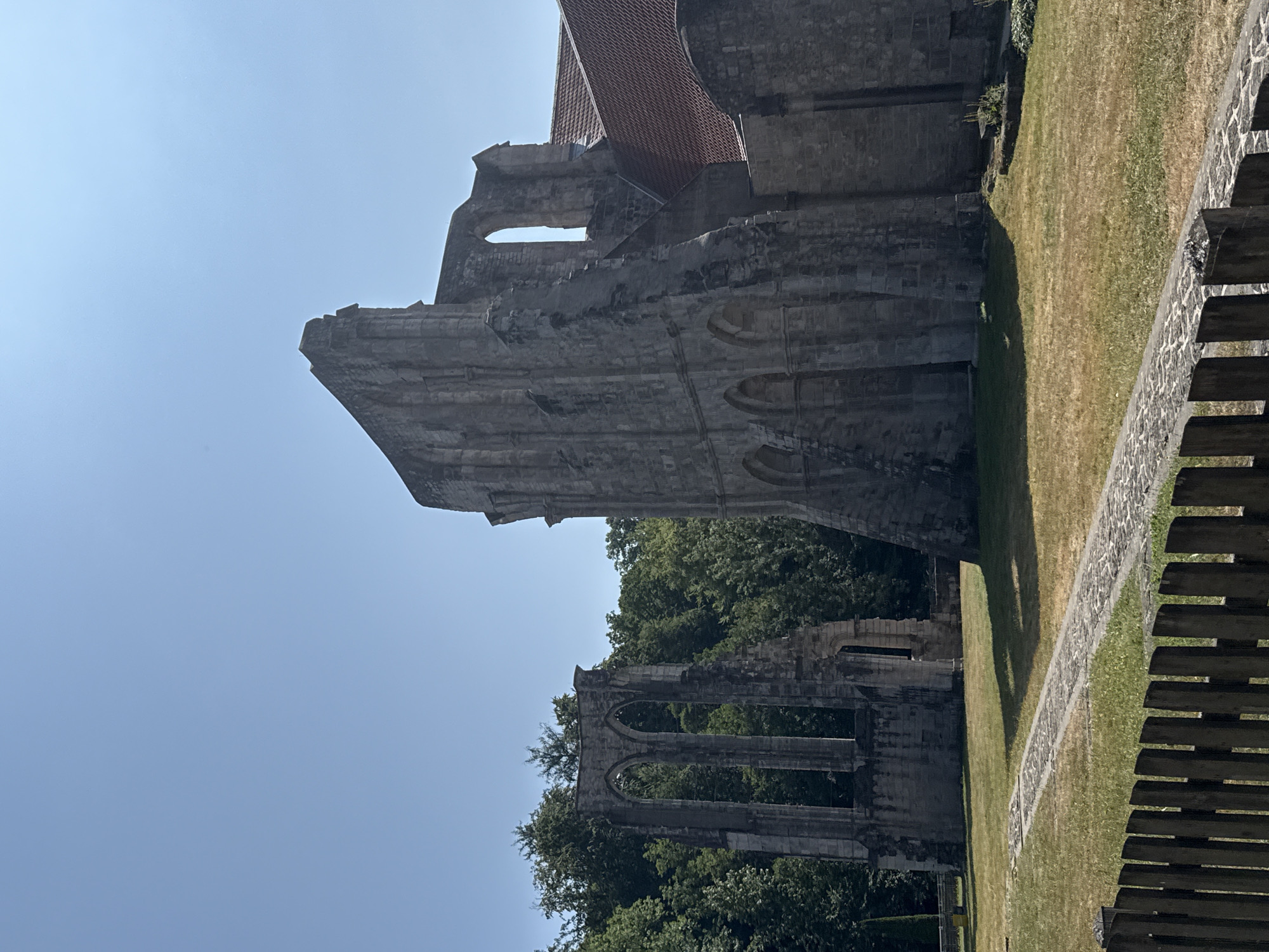The image depicts the exterior of the Walkenried Abbey in Walkenried, Germany, on a clear day. The foreground features a rustic wooden fence. Beyond the fence, a stone pathway meanders across a lawn dotted with dry grass. To the left of the pathway, ancient stone ruins, characterized by arched windows and columns, stand against a backdrop of lush green trees. To the right, a portion of the abbey's intact structure is visible, with a red-tiled roof. The sky is a clear, bright blue. There are no people or animals present in the scene.