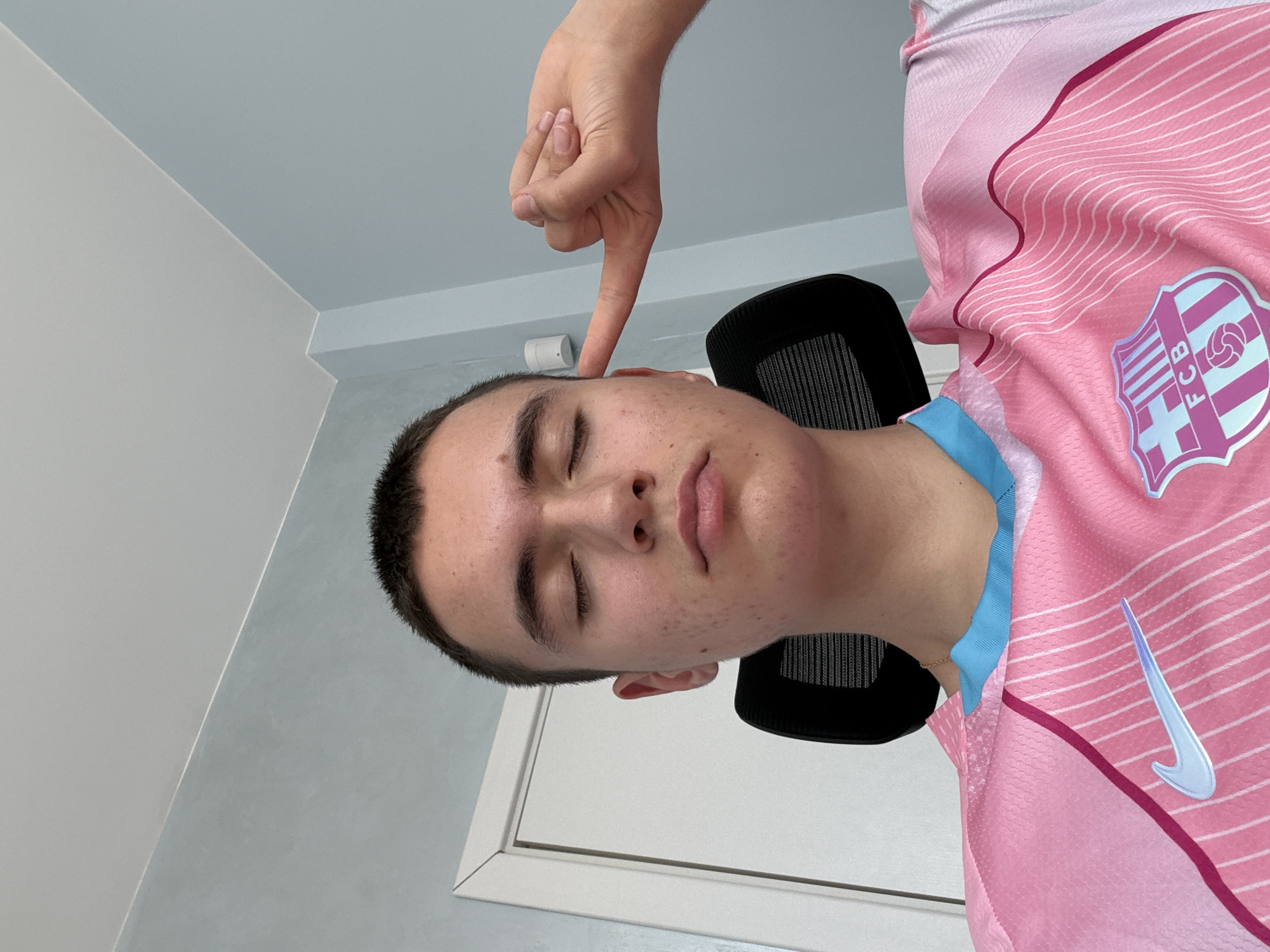 A young male, appearing to be in his late teens or early twenties, is depicted in an indoor setting, described as being located in Sofia, Bulgaria. He is seated, with his eyes closed and a relaxed facial expression, featuring slightly parted lips. His short, dark, closely cropped hair is visible. He wears a pink short-sleeved FC Barcelona athletic jersey, which includes a light blue collar accent. The Nike swoosh logo is visible on his right chest, and the FC Barcelona crest is on his left chest. His right hand, positioned on the viewer's left side of the frame, has its index finger extended and pointing towards his right temple/ear area, with the other fingers curled into a loose fist. Behind him, the black mesh backrest of a chair is partially visible. The room features light blue-grey walls and a white ceiling. A white door frame is visible on the left side of the image, and a small white cylindrical object, possibly a sensor or fixture, is mounted on the wall to the right of the subject's head. The lighting appears even and diffused.