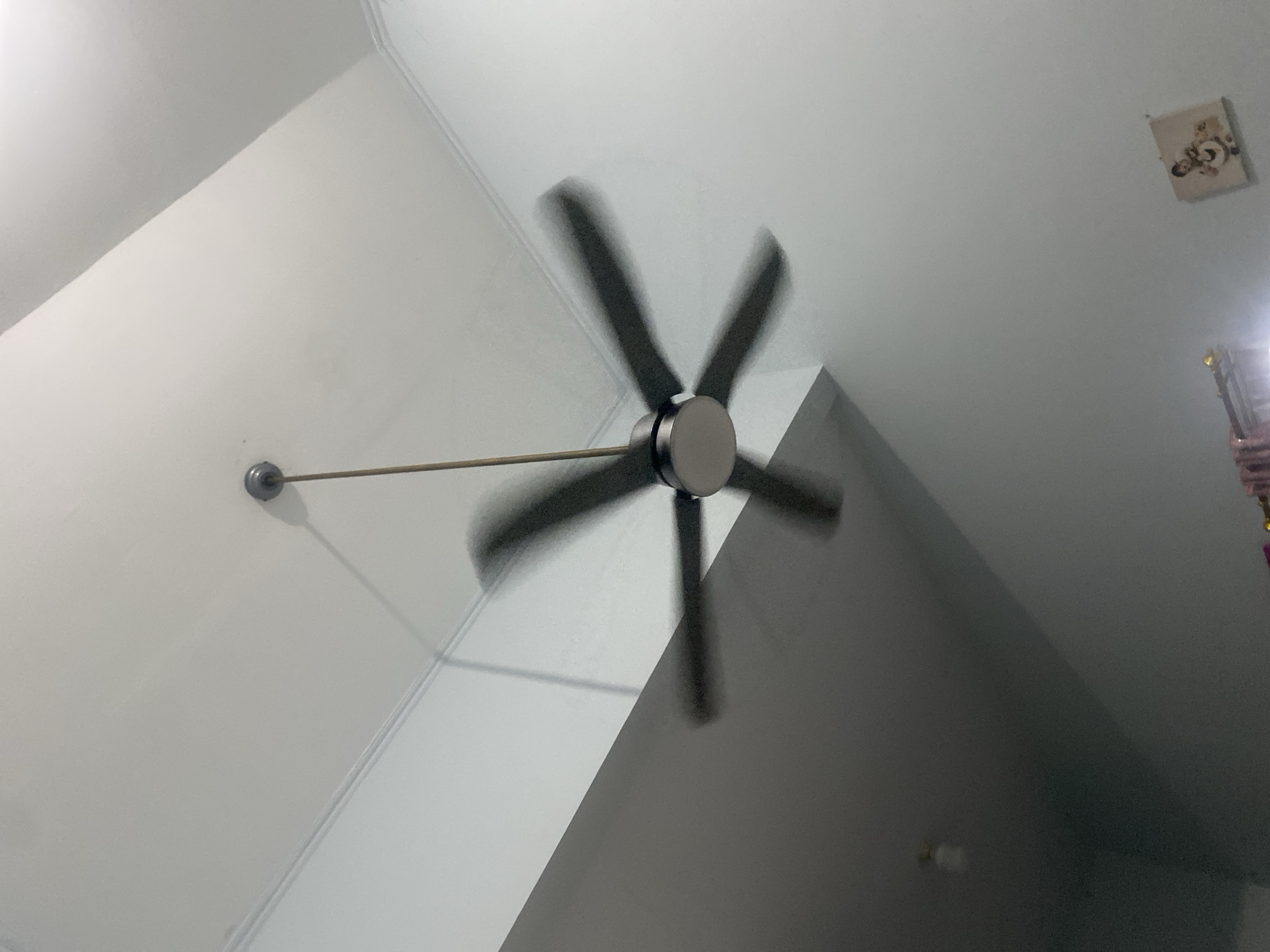 A ceiling fan with dark blades is captured in motion, creating a blur effect. The fan is suspended from the ceiling by a thin metal rod. The ceiling itself is white and features ornate molding along the edges. A portion of a wall, painted a light color, is visible on the right side of the image. The environment appears to be indoors, possibly a residential setting, in the city of Dukuh, Indonesia. The lighting suggests it may be daytime, but the indoor setting and the presence of artificial light sources are also possible. There is a framed picture on the wall on the right side.