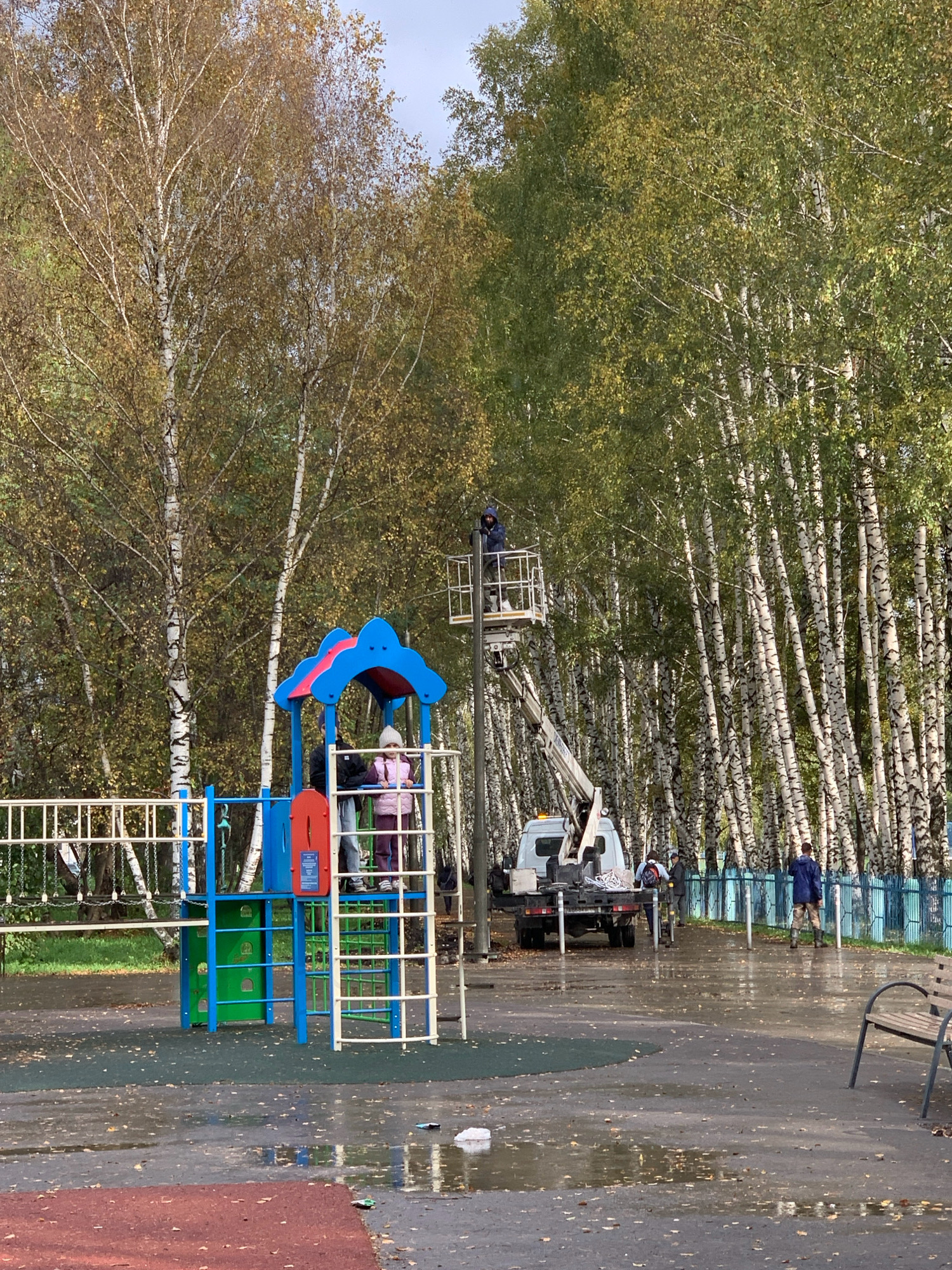 The image depicts an outdoor scene, likely a playground, in Prokopyevsk, Russia, during autumn. The ground is wet and reflects the surroundings, indicating recent rain or damp conditions. Fallen leaves are scattered across the paved surface. In the background, a dense stand of birch trees with white trunks and partially yellowed foliage dominates the landscape.
In the foreground, a brightly colored children's playground structure, featuring red, blue, and green elements, is visible. Several children and adults are present. Two children, one wearing a pink puffer jacket and a light-colored hat, and another partially obscured by a jacket, are on the playground structure.
Further back, a white maintenance truck with a hydraulic lift is parked. A person is positioned in the lift basket, appearing to be working on a pole or overhead structure. Several other individuals, presumably workers, are standing near the truck and on the ground. A blue metal fence runs along the edge of the area. A wooden park bench is situated to the right of the image. The overall atmosphere suggests a public space undergoing maintenance or activity.