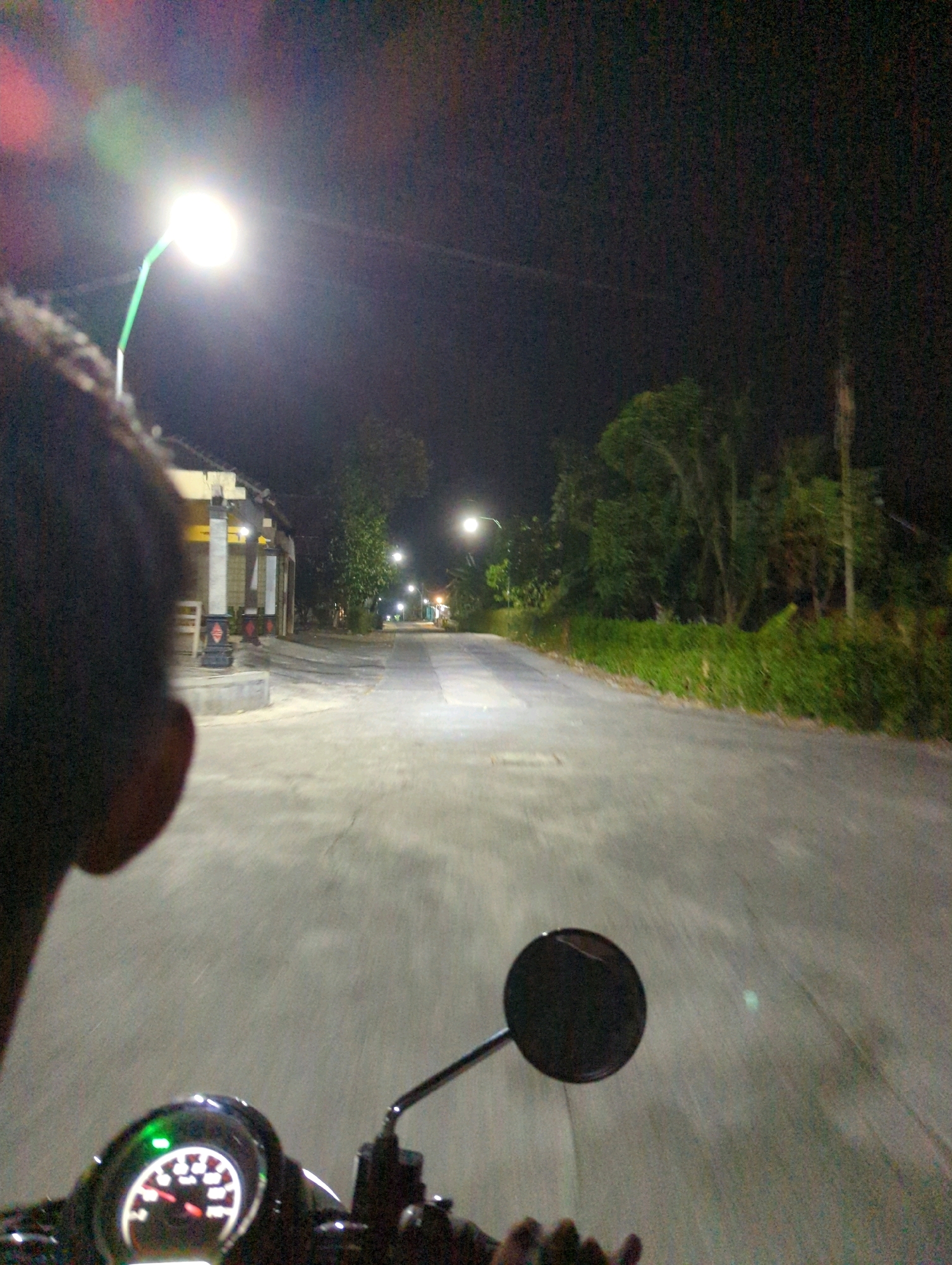 The image depicts a nighttime scene on a road in Kunti, Indonesia. In the foreground, a portion of a person's head is visible on the left, out of focus. The viewpoint is from the perspective of someone riding a motorcycle, as evidenced by the handlebars and a dashboard with a lit green indicator and a speedometer visible at the bottom. The road stretches ahead, illuminated by streetlights positioned on the left side of the road. On the left, there are buildings with some lights on, possibly shops or houses. Trees and dense vegetation line the right side of the road, creating a natural barrier. The sky is dark, indicating it is late at night. The overall atmosphere is one of a quiet, rural road at night.