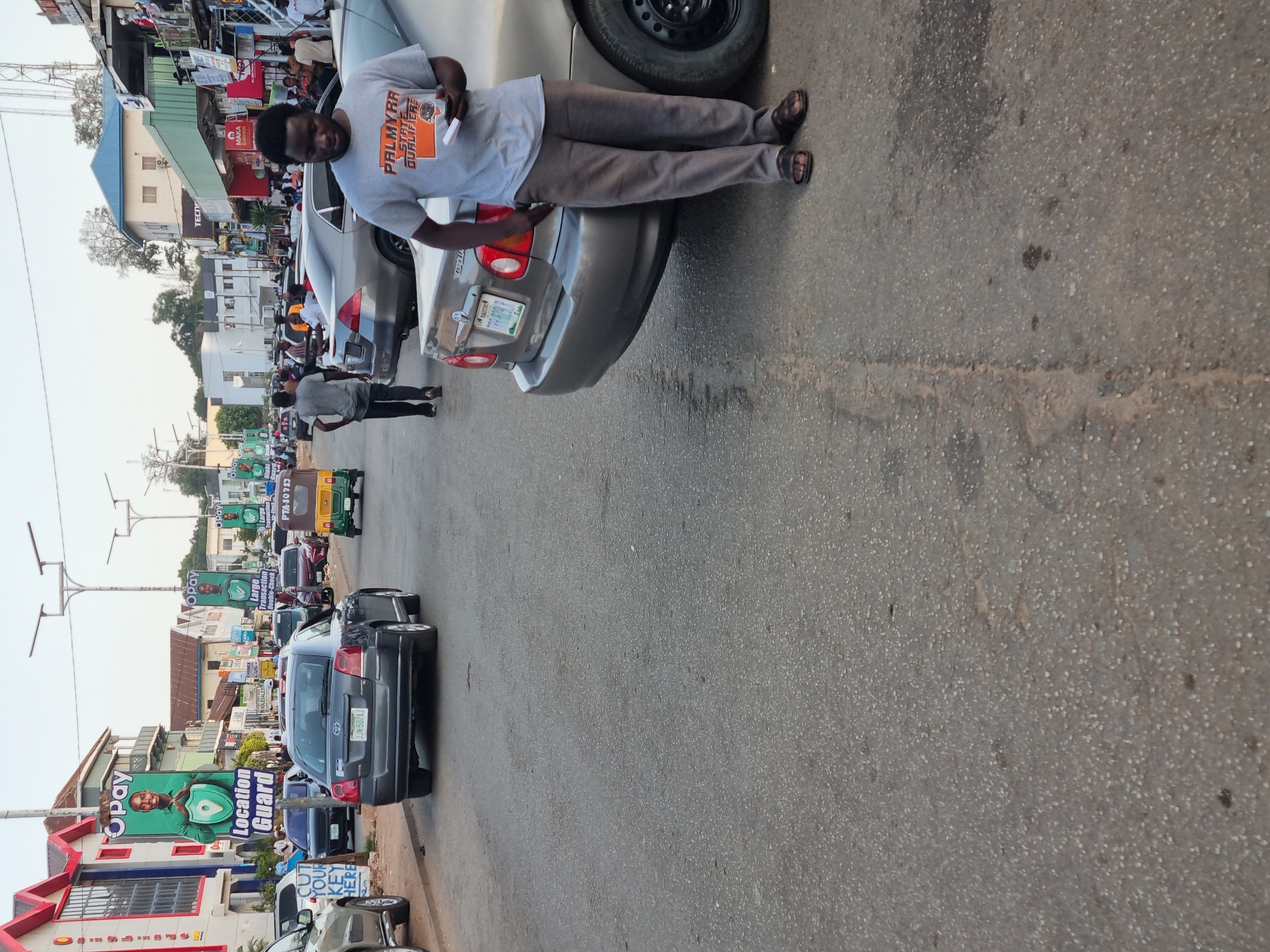 The image shows a busy street scene in what appears to be the city of Jos, Nigeria. The image shows a man wearing a grey t-shirt and grey pants standing beside the rear of a silver car. He is holding an object in his hands. The license plate of the car reads "TRT-300DL." Several other cars are parked along the street, which is lined with buildings, shops, and advertisements. Some of the businesses advertised include "OPay," "Location Guard," "CUT YOUR KEY HERE" and "MAKA." People are walking on the street and near the parked vehicles.