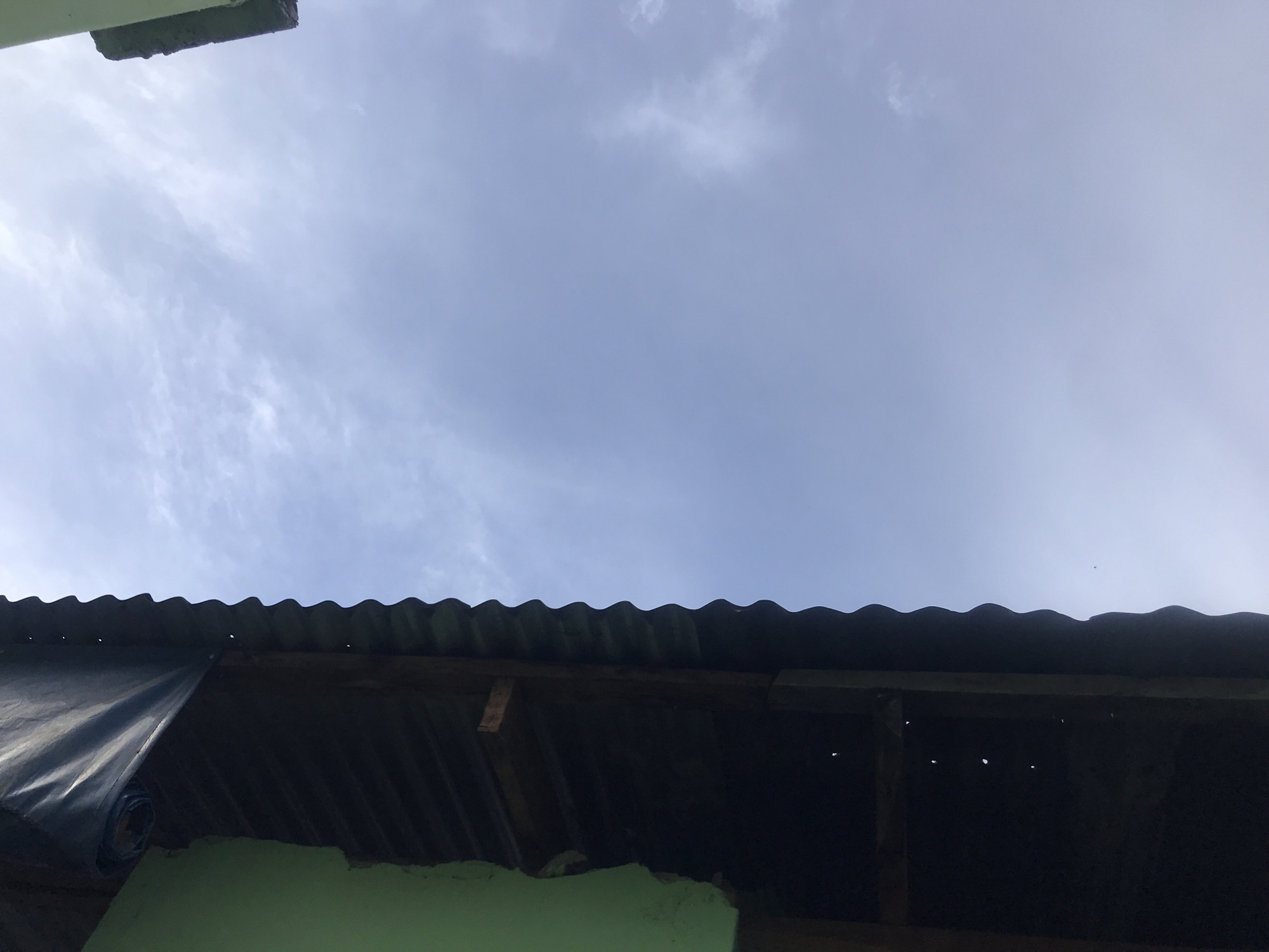 The image, taken in New Stadium, Nigeria, shows a close-up, low-angle view of a corrugated metal roof extending from a green-painted structure. A dark tarp is partially unrolled and draped over the edge of the roof. The sky, mostly overcast with light clouds, occupies the right two-thirds of the frame. Some wooden beams are visible beneath the roof.
