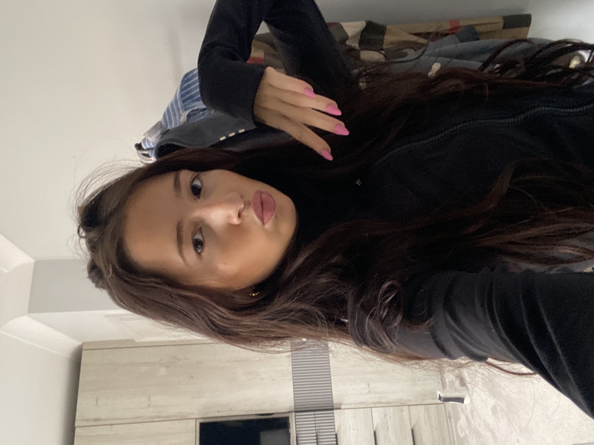 A young woman with long brown hair, light skin, and pink fingernails poses for a selfie. She is wearing a black turtleneck top and is lying on a bed with a blue and white striped shirt and patterned blanket visible in the background. The location appears to be an interior space with light-colored walls and cabinets. It cannot be determined from the image if the location is within Welwyn Hatfield, United Kingdom.