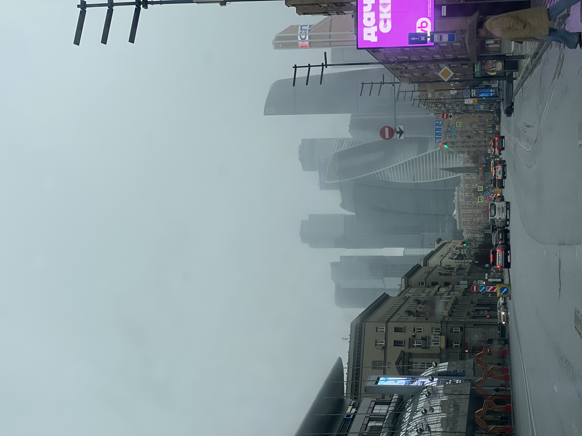 The image depicts a street in Moscow, Russia on a foggy day. Tall, modern skyscrapers of the Moscow International Business Center (Moscow City) are visible in the background, obscured by the fog. A street with traffic flows along the right side of the image. There are cars, vans, and potentially a bus visible in the traffic. Buildings line the street on both sides, with older architectural styles closer to the viewer. Street signs are visible along the road. A large billboard with a pink advertisement stands out on the right. A person in a coat and hat is walking along the right sidewalk with an umbrella.