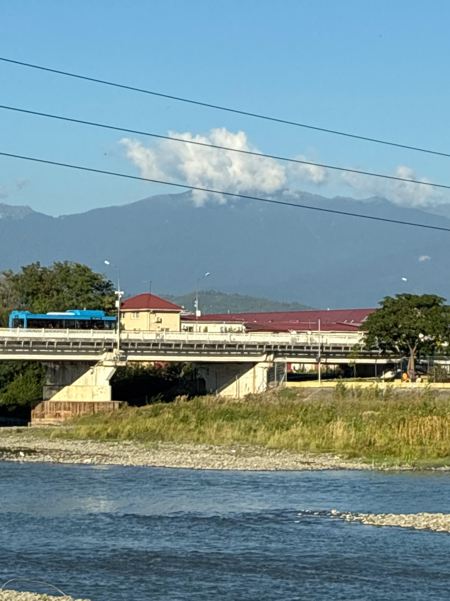 A blue trolleybus is moving across a concrete bridge, its windows reflecting the bright blue sky. The bridge spans a wide, rocky river, its waters shimmering with sunlight. On the other side of the bridge, buildings with red roofs are visible, nestled among lush green trees. In the background, a range of majestic mountains rises against the clear sky, with a few fluffy white clouds drifting near their peaks. Power lines crisscross the sky above the scene, adding a touch of human infrastructure to the natural landscape. The overall scene is peaceful and serene, with a sense of quiet movement and natural beauty. Based on the architecture and the landscape, this image was likely taken in or near the city of Sochi, Russia, potentially along the Mzymta River or a similar waterway that flows through the region.