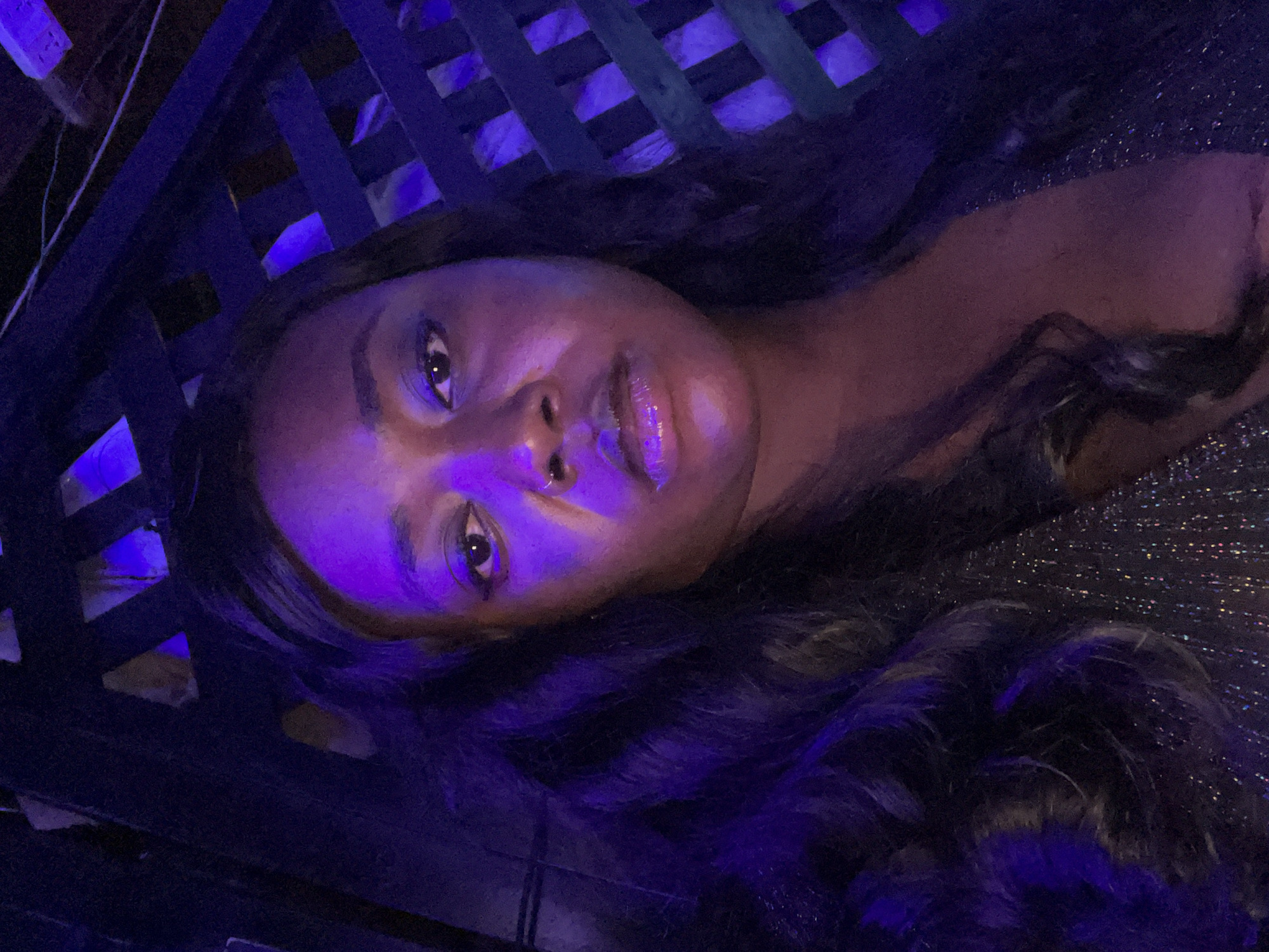 The image presents a headshot-style portrait of an adult female with dark skin, directly facing the camera. She has long, dark, wavy hair draped over her shoulders and chest. A small, metallic nose ring is visible on her left nostril. Her lips appear to be coated with a reflective or glossy substance. She is wearing a dark garment that exhibits a subtle sparkle or shimmer, consistent with sequins or glitter, and features a V-neckline.
The scene is dominated by strong blue and purple ambient lighting, which illuminates portions of her face and hair while creating deep shadows elsewhere, particularly around her eyes and under her chin. The background is dark and appears to feature a wooden lattice or trellis structure, through which additional blue and purple light sources are visible, suggesting decorative lighting within a dimly lit environment. There is no visual information within the image to confirm the location is the city of Abuja, Nigeria.