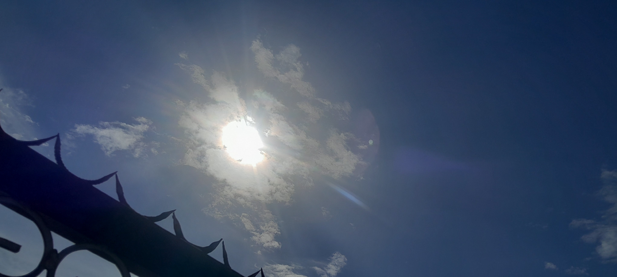 The image depicts a bright sun obscured by clouds, with visible sun rays emanating outwards. In the foreground, a dark, spiked metal fence or gate occupies the lower left portion of the frame, silhouetted against the sky. The sky is a deep blue, with scattered white clouds. There are no people or animals visible in the image. The scene appears to be an outdoor view looking upwards, likely in Gura Top, Nigeria.
