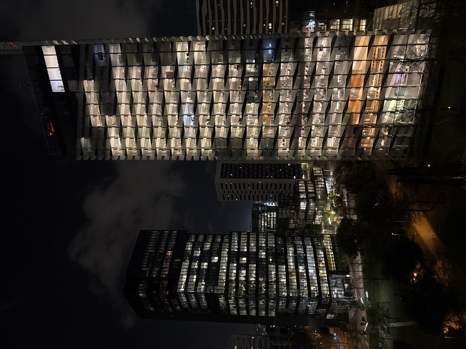 At night, tall buildings are illuminated, creating a grid-like pattern of light and shadow. The architecture features unique exterior designs, with angled and protruding elements on some buildings. Dark clouds hang in the sky, partially obscuring the structures. Paved areas and dark green spaces stretch beside the buildings, hinting at an urban setting. Distant buildings stand behind the illuminated towers. The image is rotated for a slightly disorienting perspective.