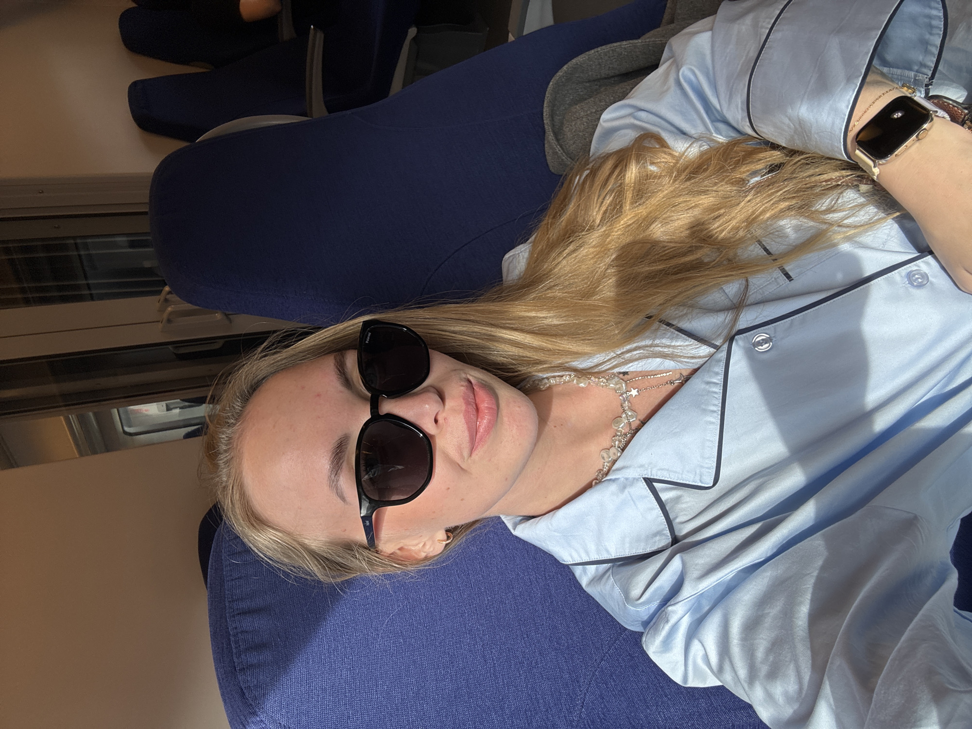 A blonde woman wearing sunglasses and a light blue pajama-style top is reclining on a dark blue cushioned seat. The setting appears to be inside a train car, as evidenced by the window in the background. There are other seats visible behind her. No location information relating to Sochi, Russia, is visible.