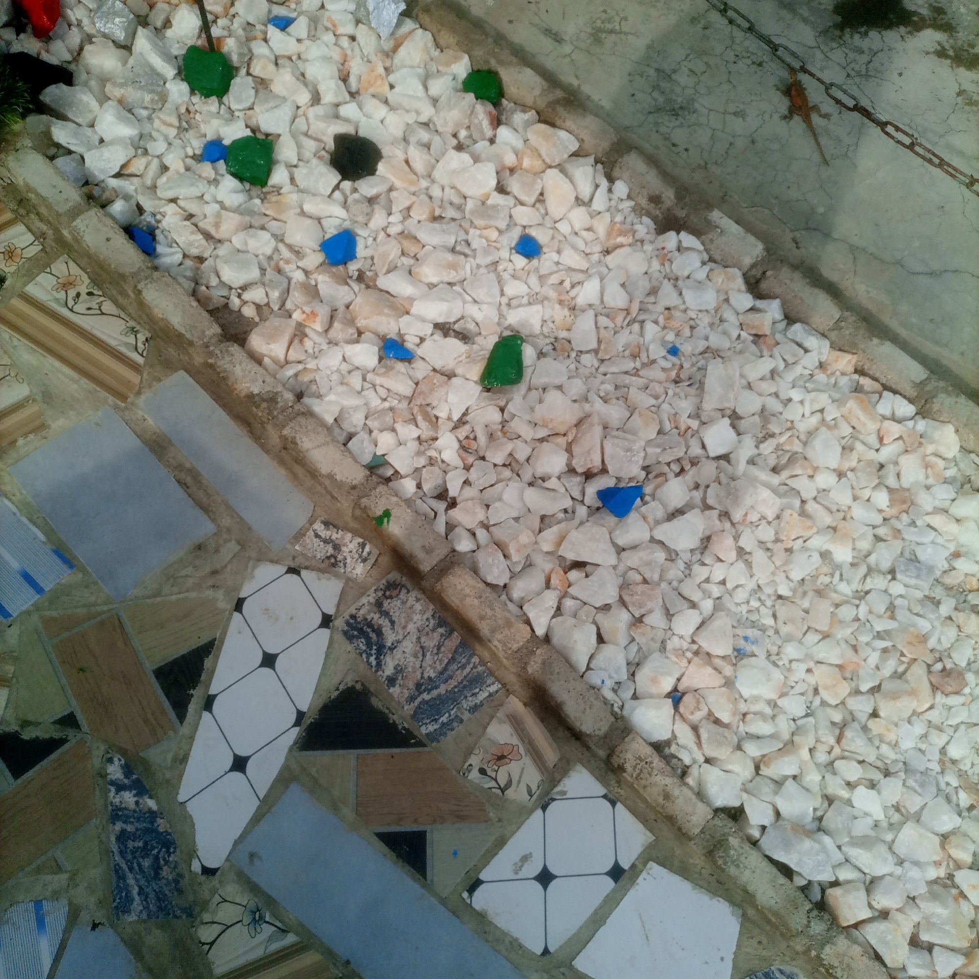 The image shows a mosaic pathway bordering a garden bed filled with white gravel and small, colorful stones. The mosaic is constructed from broken tiles of various patterns and colors, including geometric designs and floral motifs, set into cement. The gravel bed contains scattered green and blue stones. In the background, a concrete surface is visible, cracked in places, with a metal chain and a small lizard. The overall scene suggests an outdoor decorative area, possibly in a residential or public space in Jalingo, Nigeria, given the style and materials.