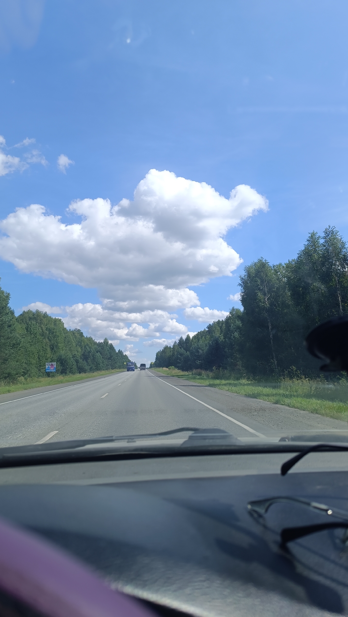 A forward-looking, eye-level, medium shot captures a view from inside a vehicle, looking out at a highway stretching through a wooded landscape under a bright, partly cloudy sky. The foreground features the dark gray dashboard of the car, with a pair of dark-rimmed glasses resting on its surface. The car is on a paved, two-lane highway with white lane markings. On either side of the road, dense green forests line the route. In the distance, several vehicles, including trucks and cars, are visible on the highway. The sky is a vibrant blue, dotted with large, fluffy white clouds. A blue and white sign is visible on the left side of the road. The overall scene suggests a journey through a rural or semi-rural area on a clear day.