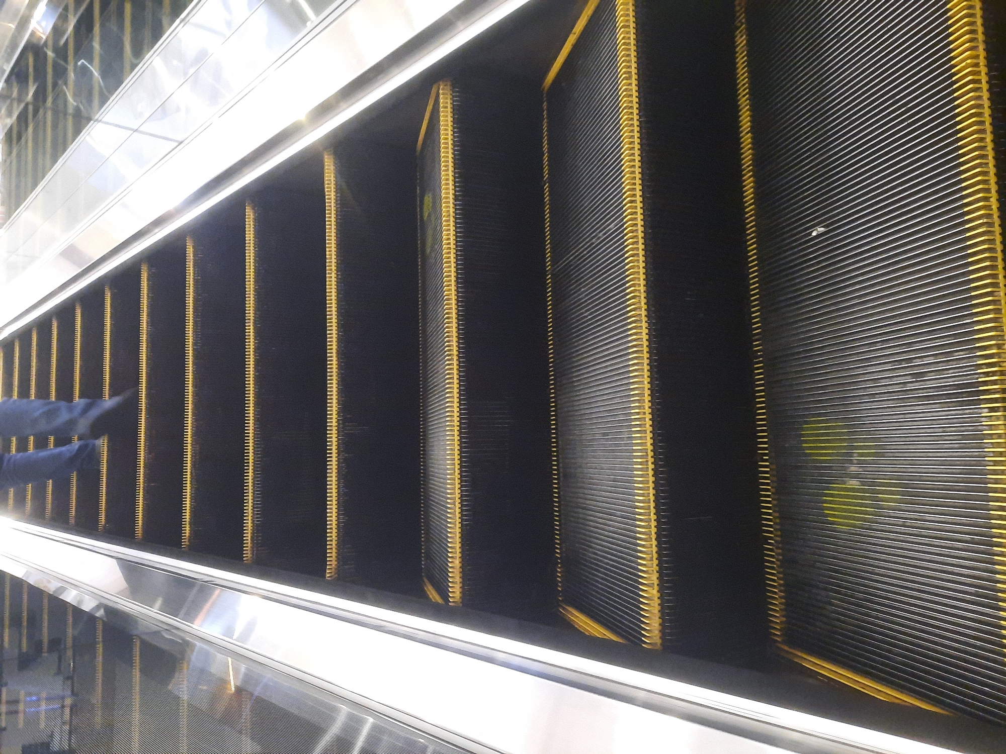 An upward-facing, eye-level shot shows a section of a black escalator with yellow trim. The steps are ridged and appear to be made of metal. The escalator railing, made of polished metal, is visible on the left side of the image, reflecting the surrounding environment. In the background, the legs of a person wearing blue jeans are visible, suggesting movement up the escalator. The overall lighting is bright, with some glare on the metal surfaces. The scene appears to be in an indoor public space, possibly a shopping mall or transportation hub, given the commonality of escalators in such locations. Without further context, the exact location within North Jakarta, Indonesia, cannot be definitively determined.