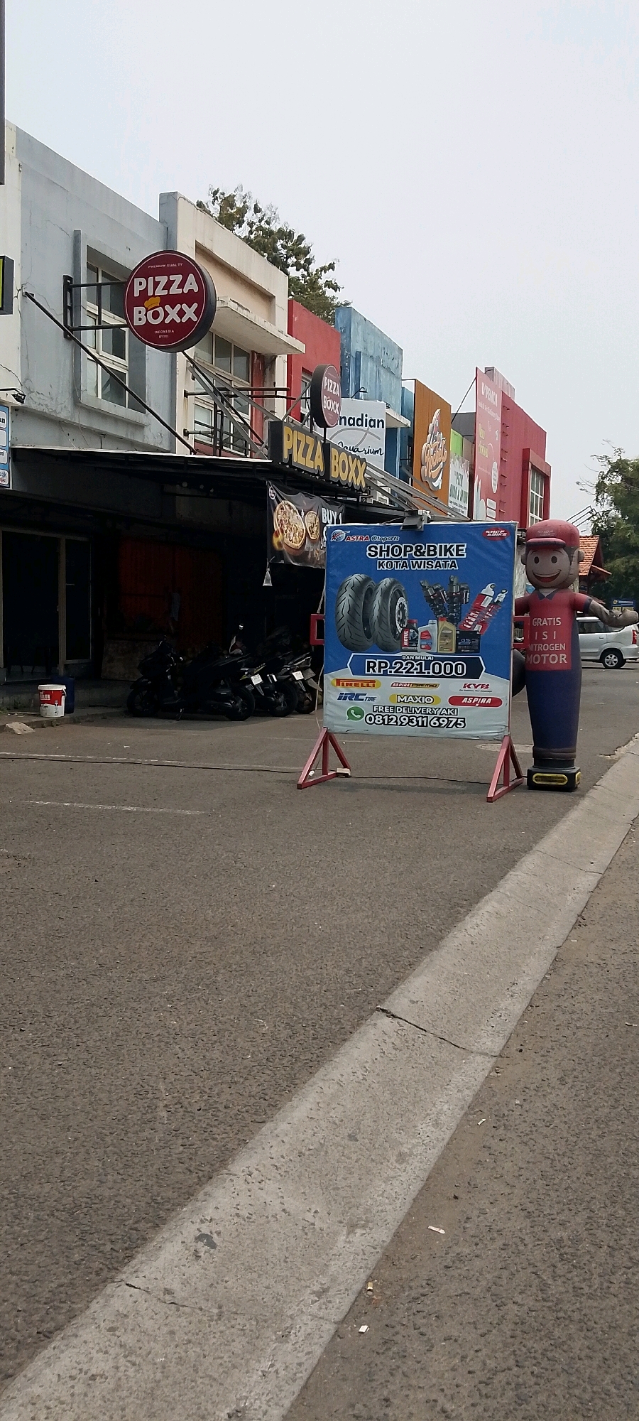 The image captures a street scene in Cileungsi, Indonesia, featuring several businesses. On the left, a building with a light gray facade prominently displays a circular red sign with "PIZZA BOXX" in white letters. Adjacent to it, another similar sign is visible. Further down the street, a banner for "SHOP&BIKE KOTA WISATA" is displayed, featuring images of tires, engine oil, and shock absorbers. A large inflatable figure of a cartoon character wearing a red cap and shirt stands next to the banner. Several dark-colored motorcycles are parked in front of a dark-colored storefront below the "PIZZA BOXX" signs. The street is paved, with a concrete curb on the right side of the image. The sky is overcast.