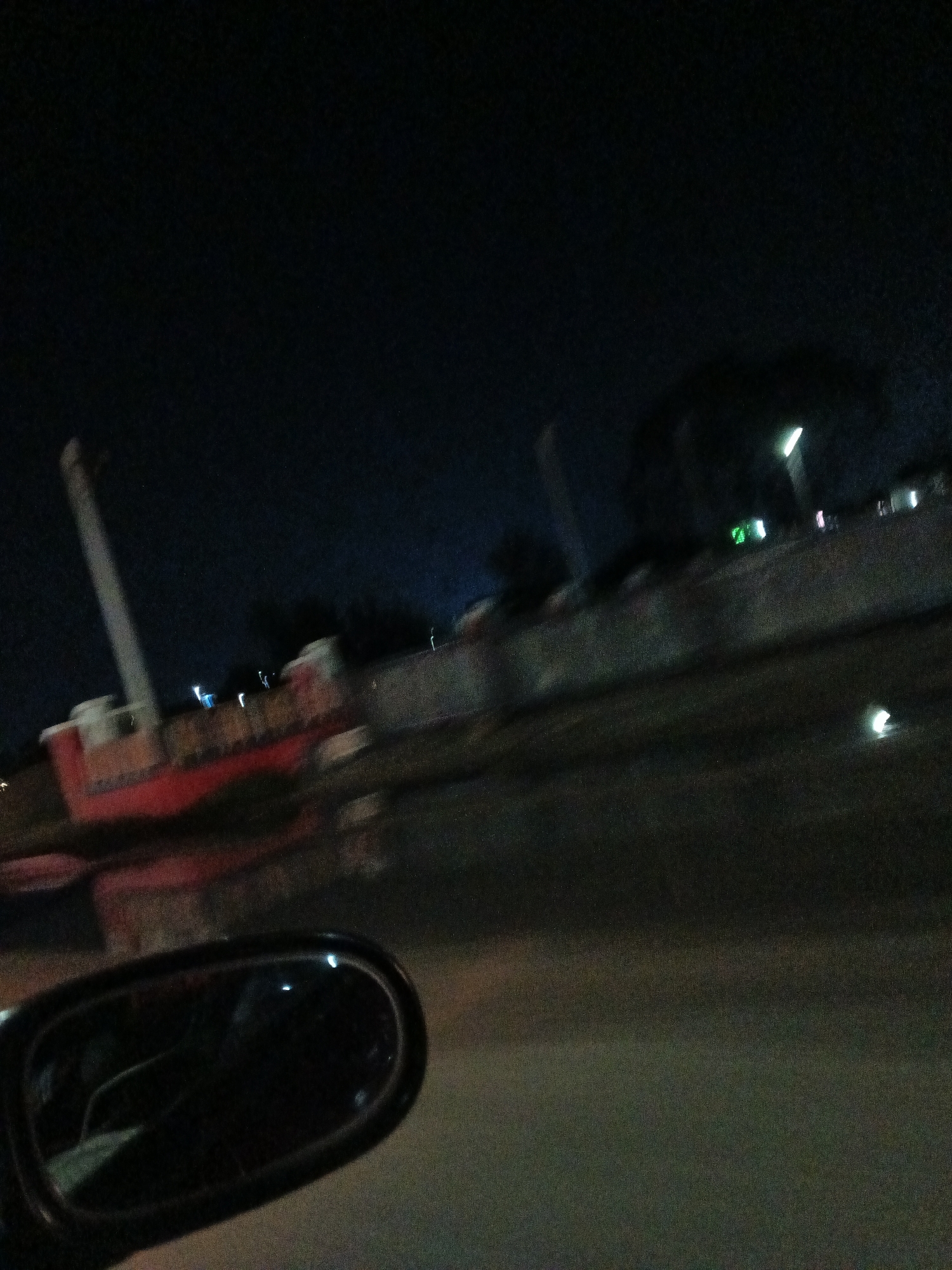The image is a blurry, dark photograph taken at night, likely from a moving vehicle. In the foreground, a car's side mirror is visible on the left. In the background, a large, red and white structure resembling a ship or a boat is partially visible, with a tall mast. There are several blurred lights scattered in the background, possibly from streetlights or buildings. The environment appears to be an outdoor, possibly urban setting at night. There is no visible indication of the city of Gura Top, Nigeria within the image.