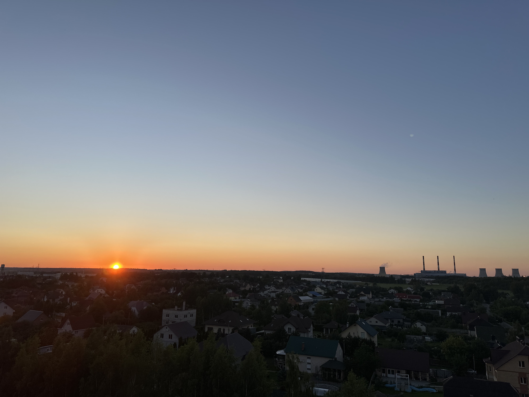 The image captures a sunset over a residential area, likely in or near Шчомысліцкі сельскі Савет, Belarus. The sun is low on the horizon, casting a warm orange glow that transitions into a pale blue sky. In the foreground, numerous houses with various roof colors are visible, surrounded by dense green trees. Further in the distance, industrial structures, including cooling towers and smokestacks emitting plumes of smoke, are silhouetted against the fading light. A faint moon or planet is visible in the upper right quadrant of the sky. The scene appears to be a panoramic view from an elevated vantage point, looking out over a mixed landscape of suburban homes and industrial facilities.