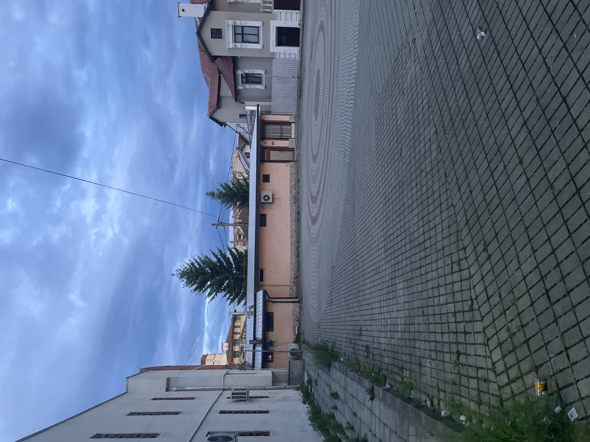 The image depicts an outdoor scene in what appears to be a town square or paved street, likely in Ihtiman, Bulgaria. The sky, occupying a significant portion of the upper frame, is cloudy with varying shades of blue and grey, suggesting an overcast day or perhaps early morning/late afternoon light. A thin power line stretches horizontally across the sky in the left half of the image.
The ground is predominantly paved with grey interlocking concrete blocks, forming a grid pattern. In the central area, a circular design composed of concentric rings of reddish-brown and grey paving stones adds a decorative element. Along the right edge of the paved area, some weeds and grass grow through the cracks, and several pieces of litter, including plastic bottles and cans, are visible.
Buildings line both sides of the paved area. On the left side, a light-colored building with several rectangular openings and two visible air conditioning units is partially seen. Further back on the left, a large coniferous tree stands prominently. To the right of the tree, a peach-colored building with a dark roof and another air conditioning unit is visible. In the background, on the right side of the frame, stands a multi-story building with a terracotta-tiled roof, light-colored walls, and detailed window frames, suggesting residential or commercial use. No people are visible, and no specific activity is taking place. There is no discernible text within the image.