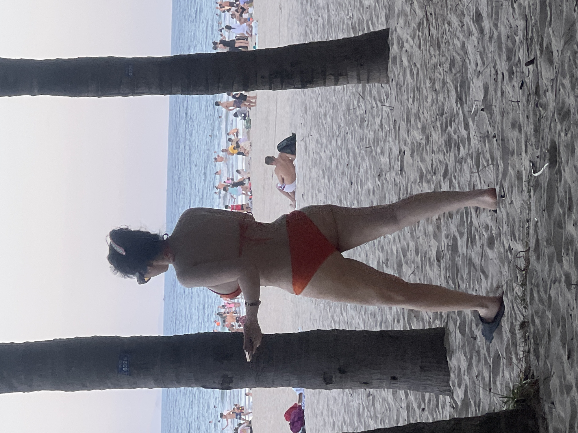 The image depicts a beach scene in Đà Nẵng, Vietnam, with the ocean and a sandy shore populated by numerous people. The weather appears to be bright and sunny, likely during the daytime. In the foreground, two palm tree trunks are visible, obscuring parts of the scene. The main subject is a woman wearing an orange bikini, positioned in the mid-ground and facing to the left. She has dark hair and is wearing sunglasses and a headband. Her left hand is holding what appears to be a cigarette.
Behind the woman, the beach is crowded with people, many of whom are in swimsuits, enjoying the ocean and the sand. The water is a light blue, indicating a calm sea. Further in the background, more beachgoers are visible, spread across the shoreline. The sand is light-colored and textured, with some debris or natural elements scattered on it. There is some visible grass growing near the foreground. On one of the palm tree trunks, there is some faint text that is difficult to read clearly but appears to be a number, possibly a marker or identification code. The overall atmosphere suggests a typical day at a popular beach destination.