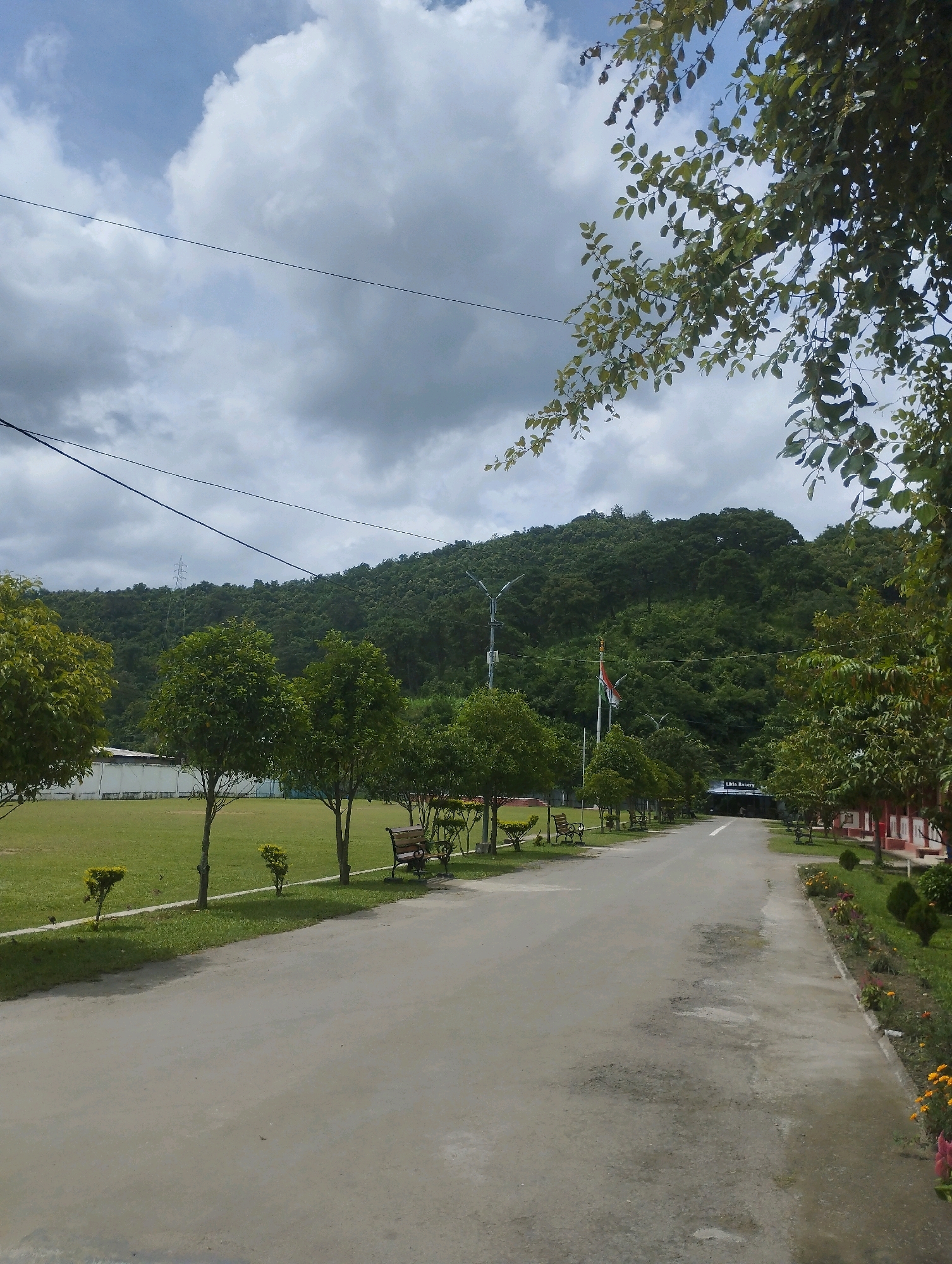 The image captures an outdoor scene in Heingang, India, characterized by a paved road leading towards a tree-covered hill under a cloudy sky. To the left of the road, a vast expanse of green grass is visible, dotted with small trees and benches. A white building is situated in the background on the left side.
On the right side of the road, a row of red-colored buildings runs along the edge, with an adjacent well-maintained garden featuring flowers and manicured bushes. The road itself is of average width, with a light gray asphalt surface showing some signs of wear and tear.
Further down the road, a flagpole stands prominently, displaying the Indian flag, which appears to be gently swaying. A sign reading "Likhita Bakery" is visible above the entrance of one of the buildings on the right. The hill in the background is densely covered with lush green trees, suggesting a natural, possibly rural or semi-rural setting within or near Heingang. The sky is overcast, with large, textured clouds dominating the upper portion of the image, indicating a day with mixed weather conditions.