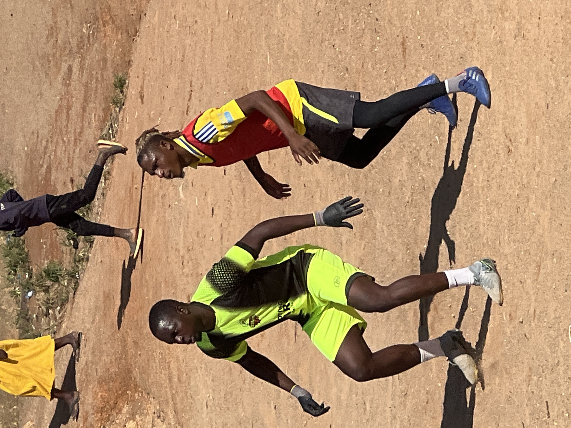 Two young men are playing soccer on a dirt field in Jos, Nigeria. The man on the left is wearing a neon yellow and black soccer jersey, neon yellow shorts, and black gloves. He is in a crouched position, looking to his right. The man on the right is wearing a yellow and red soccer bib over a yellow shirt, black shorts, and black leggings. He is looking forward and appears to be running. In the background, two other figures are walking away from the camera, one wearing a yellow garment and the other in dark clothing. The environment is dry and dusty, with sparse vegetation visible.