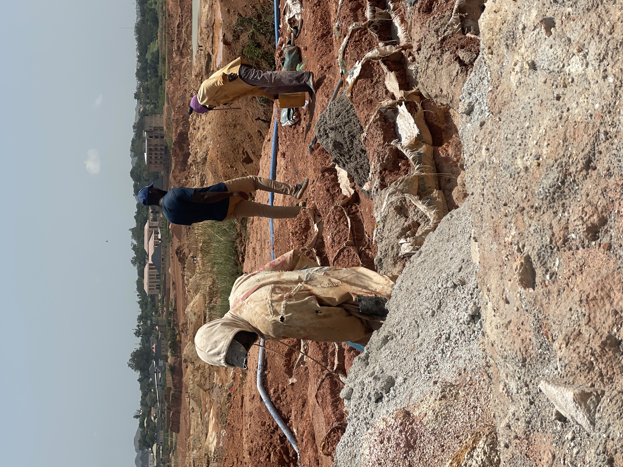 The image depicts an outdoor scene in Rayfield, Nigeria, characterized by a construction or excavation site. In the foreground, a person wearing a tan hooded garment with frayed edges and black boots is partially visible, appearing to be engaged in manual labor. Their face is obscured, but a dark material covers their head and neck. To their right and slightly behind, another person in a yellow long-sleeved shirt, brown pants, and a purple head covering is bent over, holding a yellow container and interacting with what appears to be a pile of dark material. A third person, wearing a blue cap, dark blue t-shirt, and tan pants, stands further back, observing the scene.
The environment is predominantly composed of red earth and piles of loose soil and gravel. Sacks, some appearing to be filled with material and others open, are visible, forming berms or retaining walls. Blue piping runs across the ground in the mid-ground, suggesting some form of fluid transfer. In the background, a landscape unfolds with scattered buildings, some appearing under construction or incomplete, and lush green vegetation, indicating a semi-urban or developing area. The sky is bright and clear, suggesting a sunny day. The overall impression is one of ongoing work at a site with significant earthmoving activities.