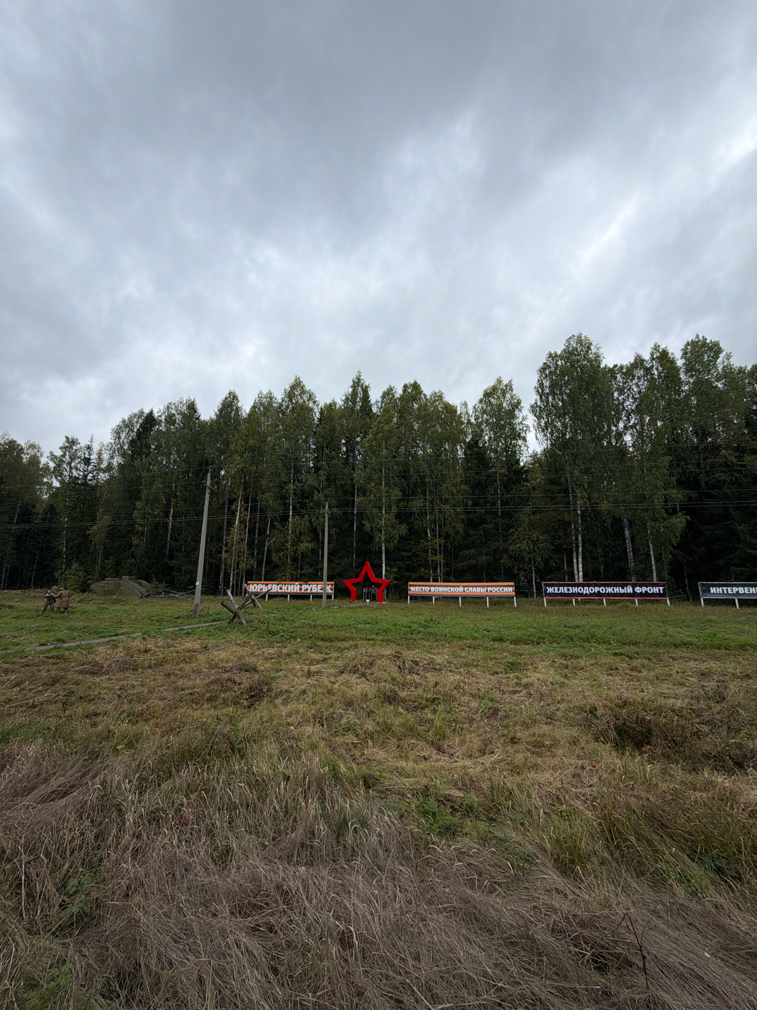 The image captures a scene in Kukhtoozerskiy, Russia, on an overcast day. A line of tall, dense green trees forms the backdrop, with a cloudy sky overhead. In the foreground, a field of dry, tall grass occupies the lower half of the frame.
On the grass, near the treeline, are several objects. A large, red, five-pointed star is prominently displayed. To its left, there are several banners with Russian text. One banner reads "ЮРЬЕВСКИЙ РУБЕЖ К" (Yuryevsky Rubesh K), another "МЕСТО ВОИНСКОЙ СЛАВЫ РОССИИ" (Place of Military Glory of Russia), and a third "ЖЕЛЕЗНОДОРОЖНЫЙ ФРОНТ" (Railway Front). To the far right, partially visible, is a banner reading "ИНТЕРВЕН" (Interven).
Further to the left, there are two figures in military-style uniforms, appearing to be static displays or mannequins. Behind them, part of a military vehicle, possibly a tank or an armored vehicle, is visible. A wooden barricade structure is also present in the middle ground. Utility poles and wires run along the edge of the treeline. The overall impression is a historical or memorial site related to military events.