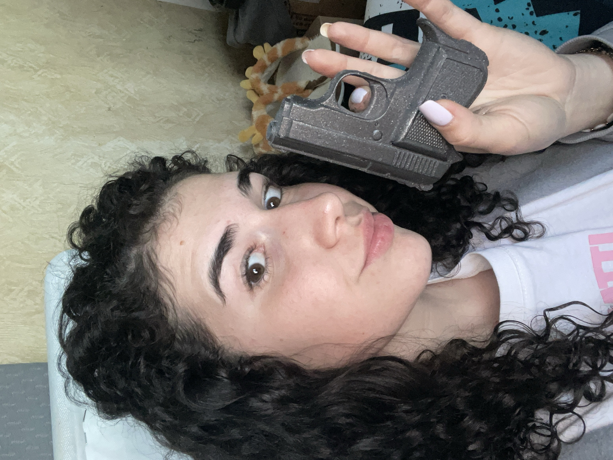 A person with dark curly hair is lying on a light-colored surface, possibly a bed or couch. They are holding a gray, possibly toy or non-functional, handgun above their head with their right hand. The person is looking up at the camera with a slightly quizzical expression. Their nails are painted a light pink color. The background includes some indistinct objects, including what appears to be a stuffed animal and a box. It is impossible to verify the location of this picture using given information.
