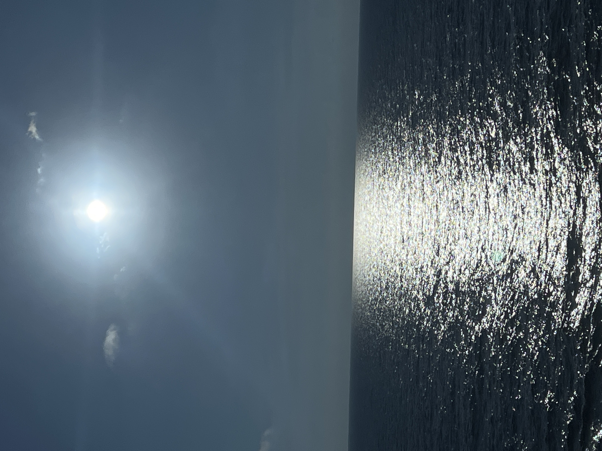 The image shows a bright sun and a cloudy sky over the ocean. A reflection of the sun shimmers on the water's surface, creating a path of light. The ocean is dark blue and appears calm, with small waves. The horizon is visible in the distance, separating the sky and the sea. There are no people or objects visible in the scene, making it a natural landscape photograph.