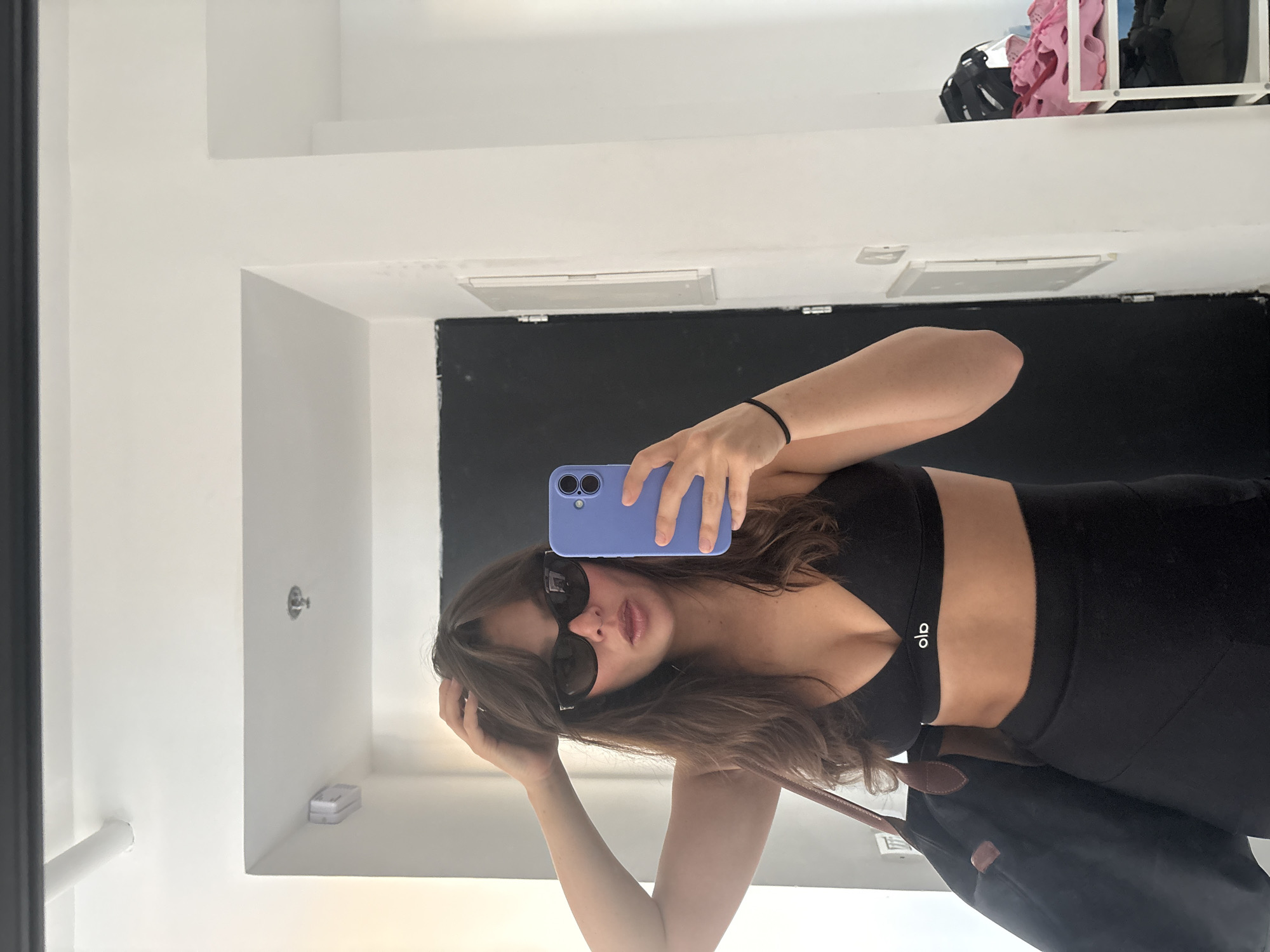 A young woman, with dark brown, wavy hair and bangs, stands in front of a mirror, taking a selfie. She is wearing a black sports bra with a small white logo, black leggings, and dark sunglasses. She holds a purple smartphone in her right hand to take the photo, and her left hand is resting on her head. A dark bag with brown straps is slung over her left shoulder. The background shows a white wall with a black door and some light fixtures. On a shelf to the right, there are pink and white shoes and a helmet. This appears to be an interior shot, possibly in a changing room or hallway. There are no visible landmarks that definitively place the location in Tel-Aviv, Israel.