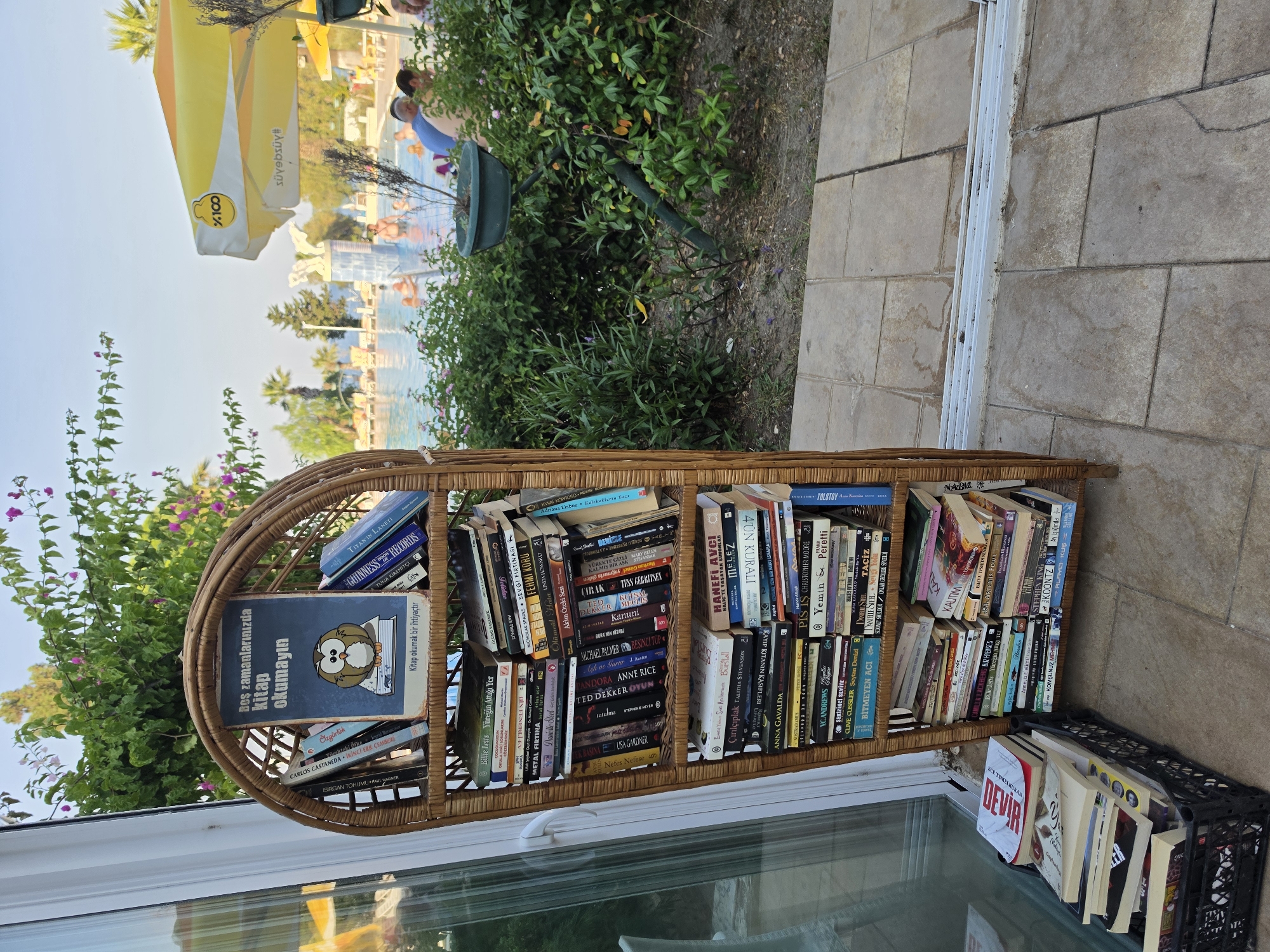 The image depicts an outdoor scene in Urla, Turkey, with a focus on a wicker bookshelf filled with books. The bookshelf is located on a paved patio area, adjacent to a swimming pool and lush greenery. In the background, people are visible enjoying the pool and relaxing under umbrellas. The sky is clear and blue, suggesting it is a daytime shot.
The wicker bookshelf is the central element, standing tall and arched, filled with a variety of books of different sizes and colors. Many of the book titles are partially visible, and some appear to be in Turkish. One book on the top shelf has a cover featuring an owl and text that reads "Boş zamanlarınızda kitap okumayın" which translates to "Don't read in your free time". Another book below it has the subtitle "Kitap okumak bir ihtiyaçtır", meaning "Reading is a necessity."
To the left of the bookshelf, a black plastic crate is filled with more books, some stacked neatly and others piled haphazardly. A glass sliding door is visible to the left of the bookshelf, reflecting some of the surroundings.
The environment surrounding the bookshelf includes green bushes and plants, some with small pink flowers. Beyond the greenery, a swimming pool with people in it and lounge chairs can be seen. Yellow umbrellas are also visible near the pool area. The paving stones on the ground are a light beige color, with some darker spots and patterns. The overall impression is one of a relaxed, holiday-like atmosphere.