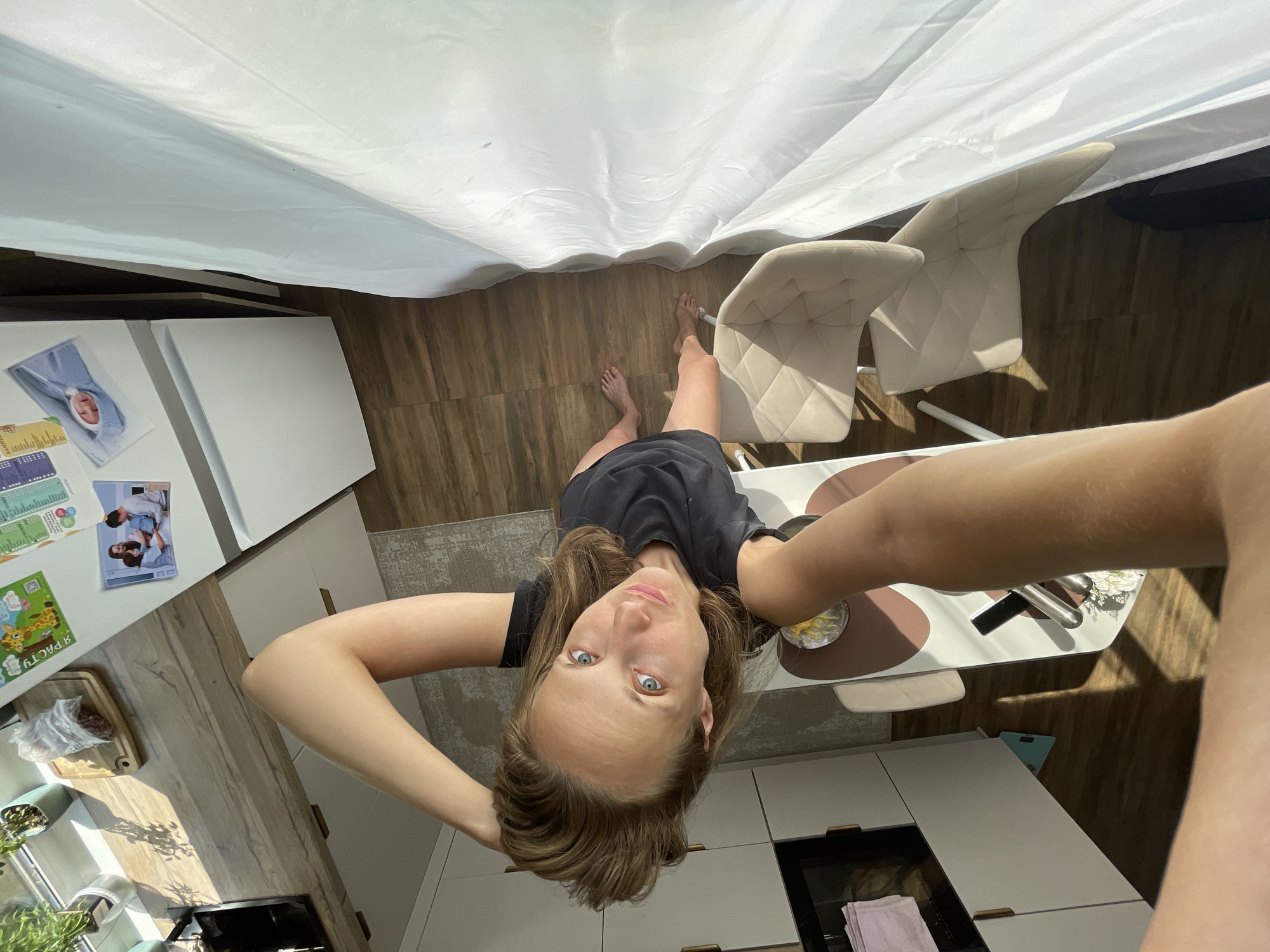 This is a first-person perspective photograph taken in a modern kitchen, likely in Подшивалово, Russia. The subject, a woman with light brown hair and blue eyes, is the central focus, positioned upside down in the frame. She is wearing a black t-shirt and appears to be holding her hair with her hands behind her head, looking directly at the camera. Her expression is neutral.
The setting includes a light-colored kitchen with wooden flooring. In the background, two light-colored dining chairs are visible. A table with a placemat and what appears to be food or a drink is also present. To the left of the subject, a white refrigerator is adorned with several colorful magnets and photographs. Among them is a magnet with a picture of a baby and another that seems to be a children's drawing. Further to the left, a countertop displays some kitchen items, including a wooden cutting board with a plastic bag of food, a plant in a green pot, and a stainless steel appliance. The scene is illuminated by bright natural light streaming in from a window, creating distinct shadows on the floor and furniture, suggesting it is daytime. A white curtain partially obscures the view, adding a soft, diffused light effect. The overall atmosphere is casual and domestic.