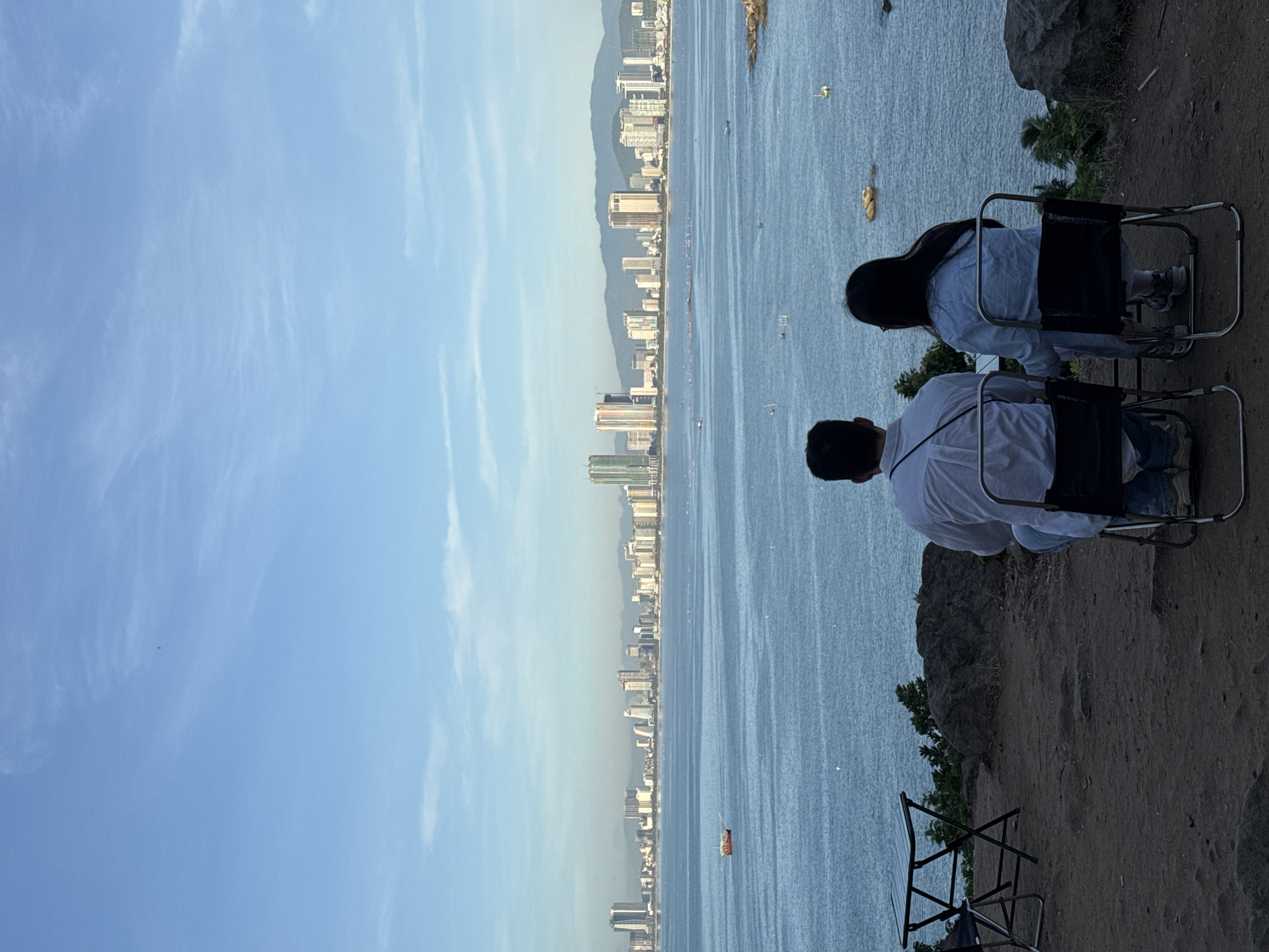 The image depicts a serene view of the city of Đà Nẵng, Vietnam, from a hilltop vantage point. In the foreground, a man and a woman are seated on folding chairs, facing away from the camera, towards the expansive sea and the city skyline. They are dressed casually, with the man in a white shirt and jeans, and the woman in a light blue denim shirt and white pants. A small folding table is positioned between them.
The sea stretches out before them, with a few boats scattered across its surface. The city skyline, a prominent feature of Đà Nẵng, is visible in the distance, characterized by a mix of modern high-rise buildings and mountainous terrain in the background. The sky is a clear, light blue, with wisps of white clouds. The ground in the foreground is a mixture of soil and rocks, with some sparse vegetation. The overall atmosphere is one of peaceful contemplation and appreciation of the scenic beauty.