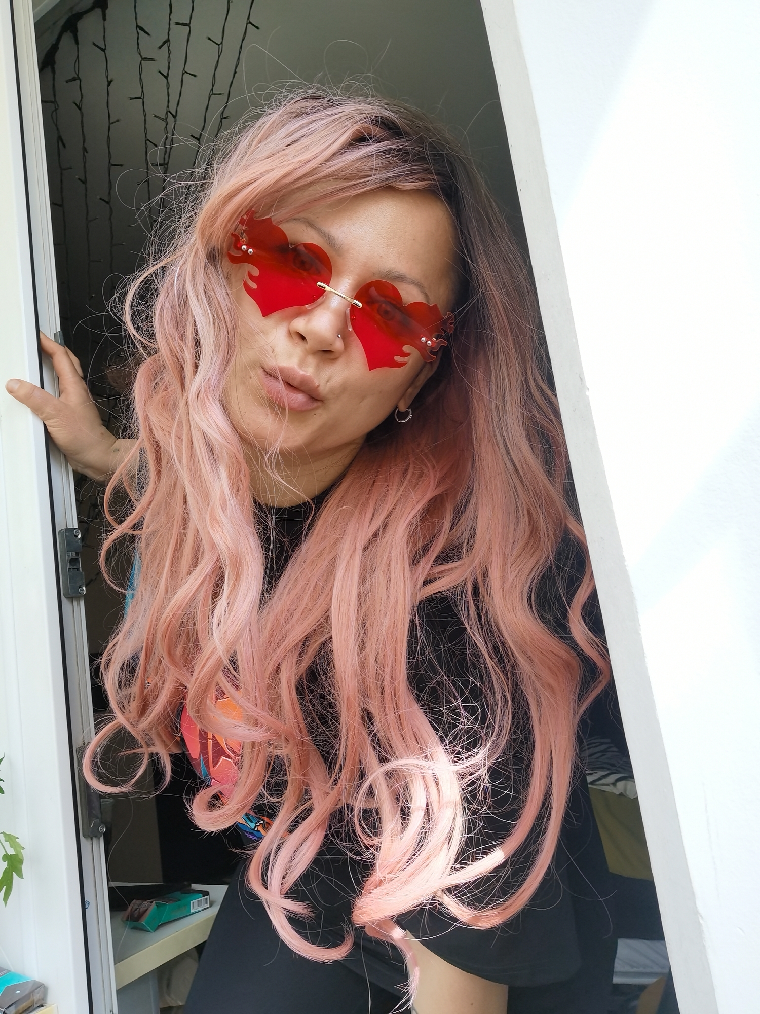 A person with pink hair and red, heart-shaped glasses is partially framed by a white doorway. The individual is wearing a black shirt with a colorful design. They are pursing their lips. Behind them, the background includes a shelf with a small package on it and what appears to be string lights hanging. The location is likely Chaikovsky, Russia.