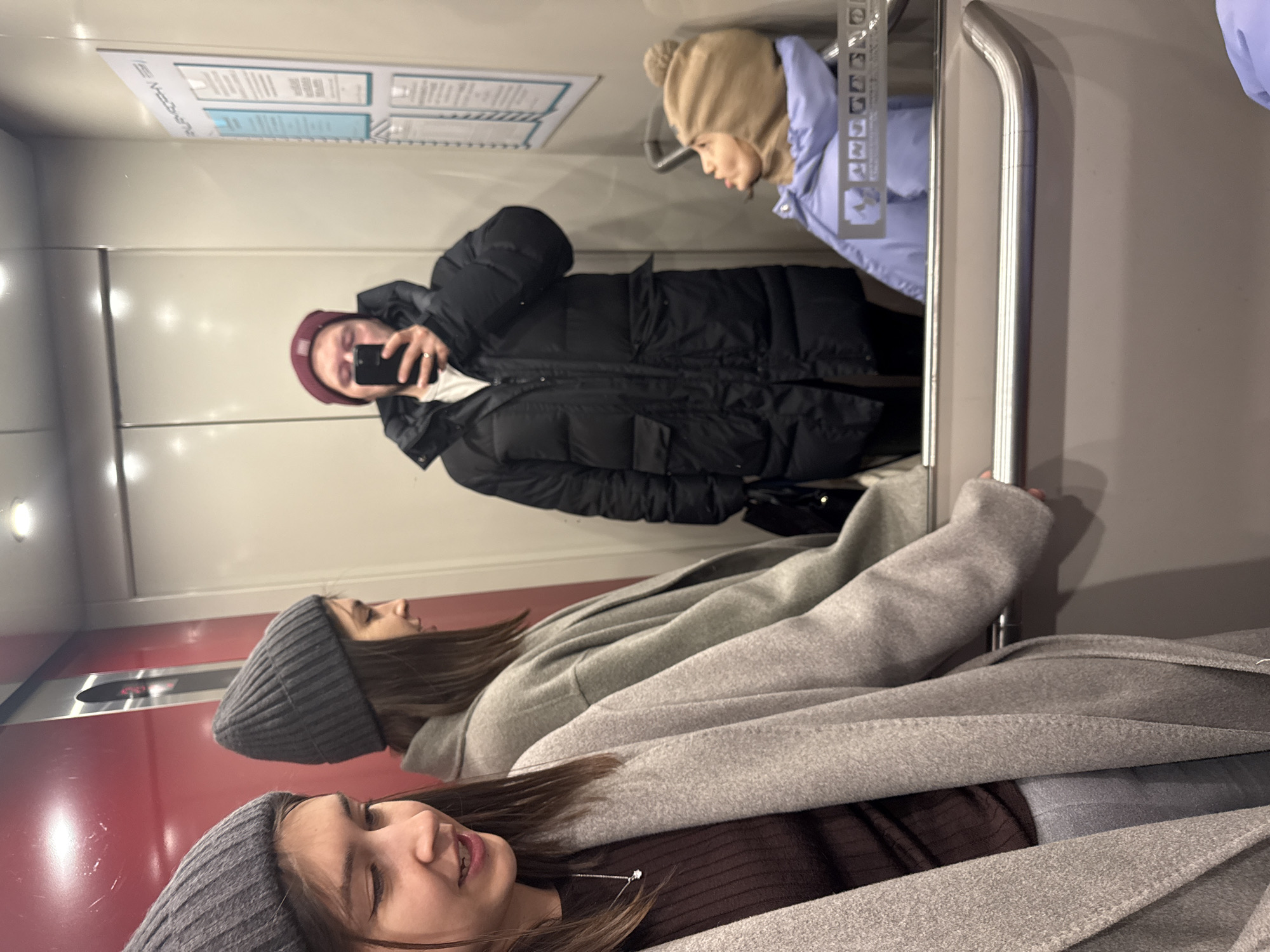 The image captures three individuals inside an elevator, primarily through a mirror reflection. The original image is rotated ninety degrees clockwise.
In the foreground, a female with long brown hair is seen from the chest up, wearing a dark gray ribbed beanie and a light gray long coat over a dark brown ribbed top. She looks upwards with a slight smile. Her reflection is visible behind her, showing her in similar attire, looking to the side.
Behind these two, and clearly visible in the mirror reflection, is a male. He wears a red or maroon beanie and a black puffer jacket over a white top. He is holding a black smartphone, suggesting he is taking a mirror selfie of the group, with the phone partially obscuring his face.
The elevator interior features light-colored walls and a large mirror. A curved silver metal handrail runs along the right side. On the wall reflected in the mirror, there is a notice board with "КРАСНОЯРСК" (Krasnoyarsk) clearly visible at the top, confirming the location as Krasnoyarsk, Russia. Below this, there are blurred lines of text or symbols. To the left, a red panel containing elevator controls or indicators is partially visible.
The individuals' winter clothing—heavy coats and beanies—suggests cold weather outside. The scene appears to be an informal moment, possibly friends or family capturing a selfie in an elevator.