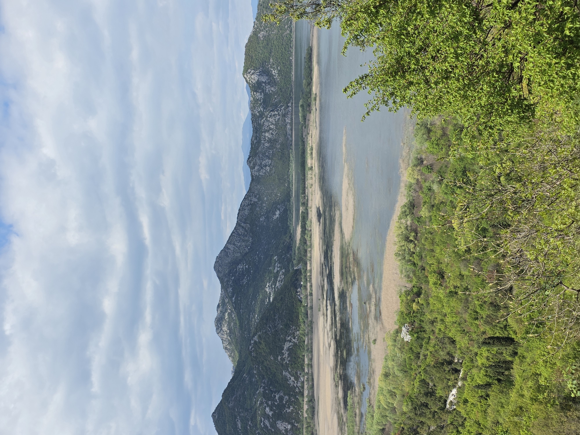 The image depicts a panoramic view of the landscape in Godinje, Montenegro. In the foreground, dense green vegetation extends along the right side of the frame, with lush trees and shrubs. A body of water, likely a lake or river, stretches horizontally across the middle of the image, reflecting the sky and surrounding mountains. The water's edge is marked by reeds and marshy areas.
In the background, a rugged mountain range rises, with rocky peaks and slopes covered in sparse greenery. The sky above is partly cloudy with blue patches. The overall scene conveys a sense of natural beauty and serenity in a rural setting.